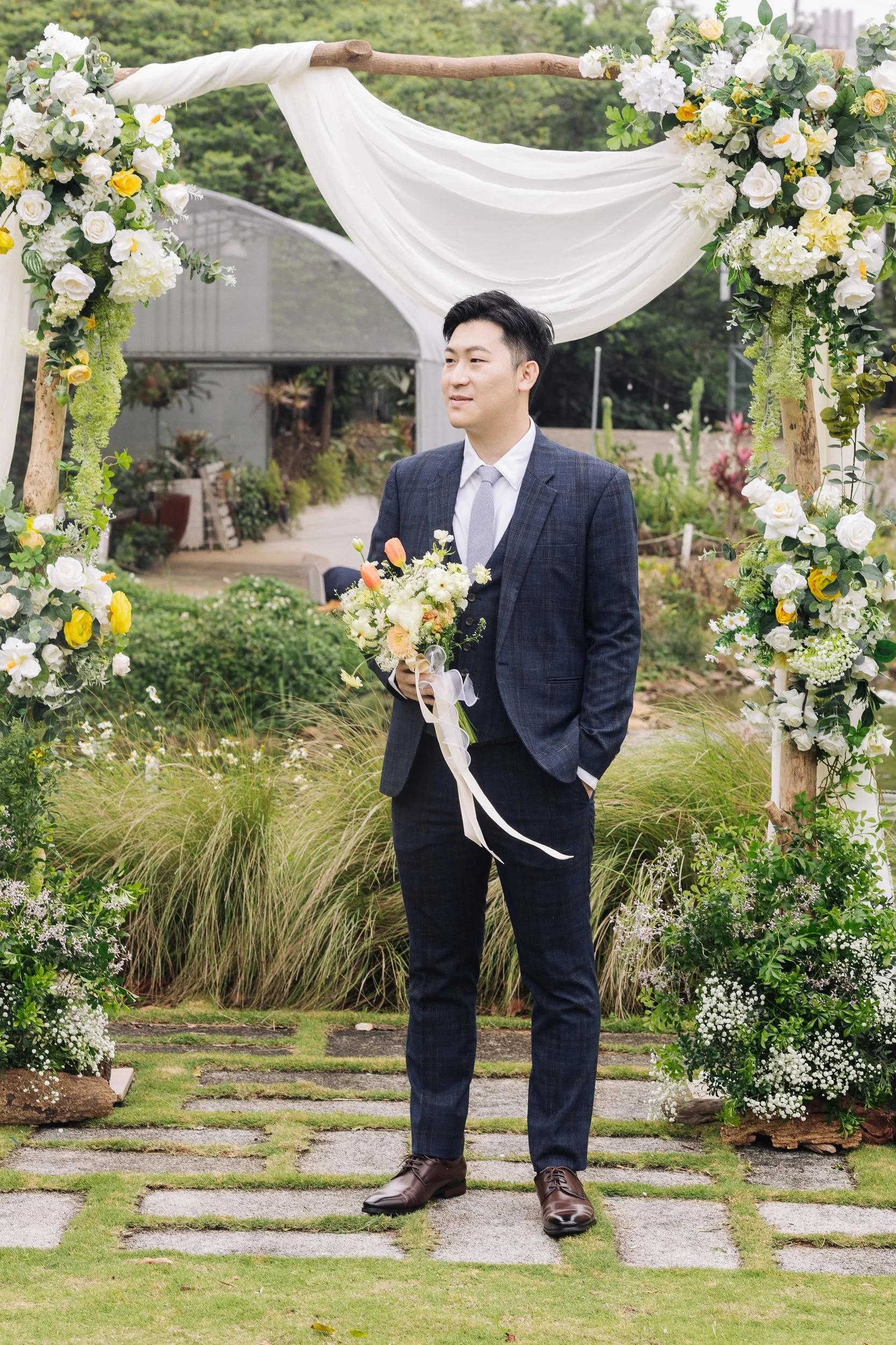 A groom in a suit holding a bouquet of flowers, standing under a floral arch at an outdoor wedding.
