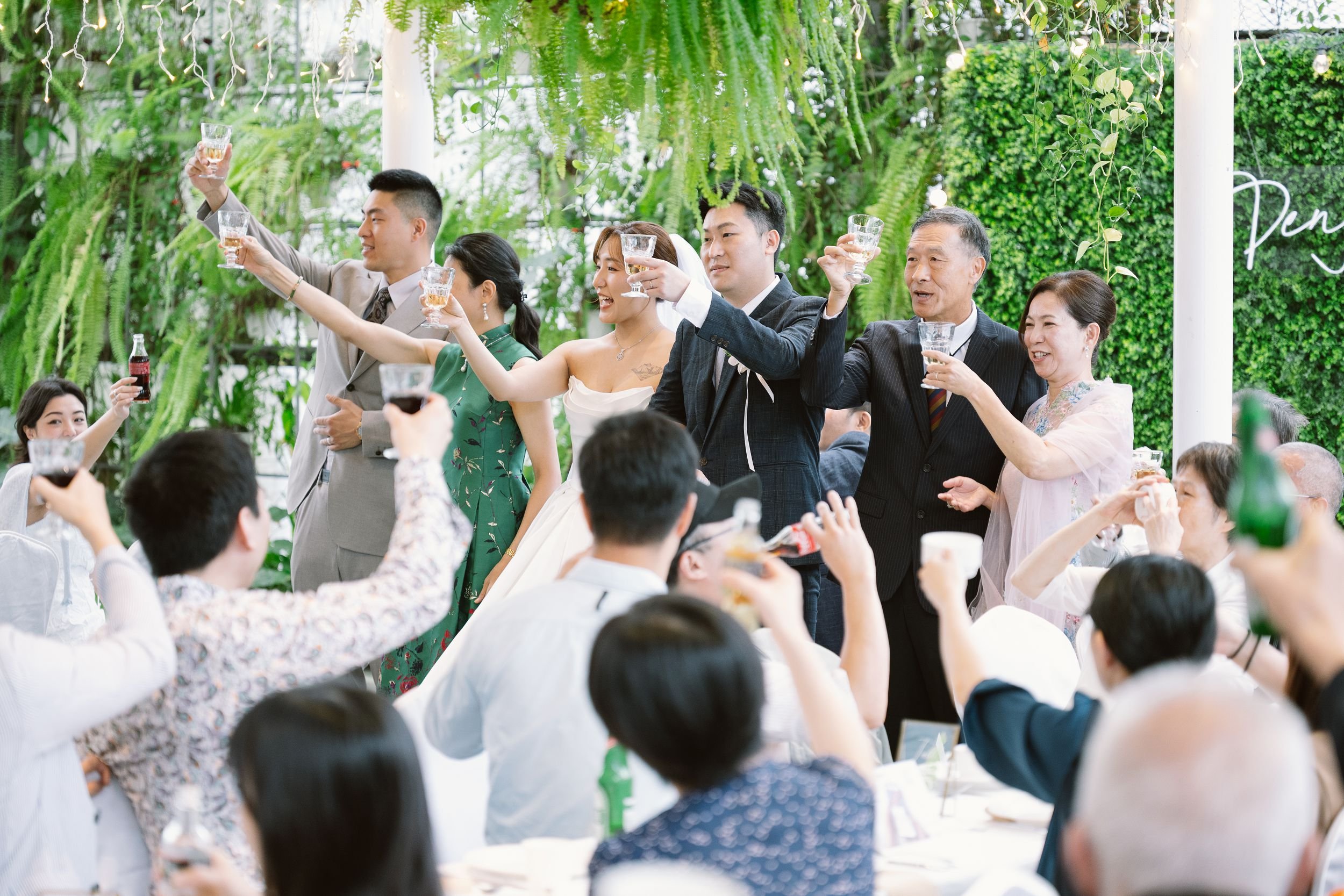 People raising glasses in a toast during a celebration in a lush green outdoor setting.