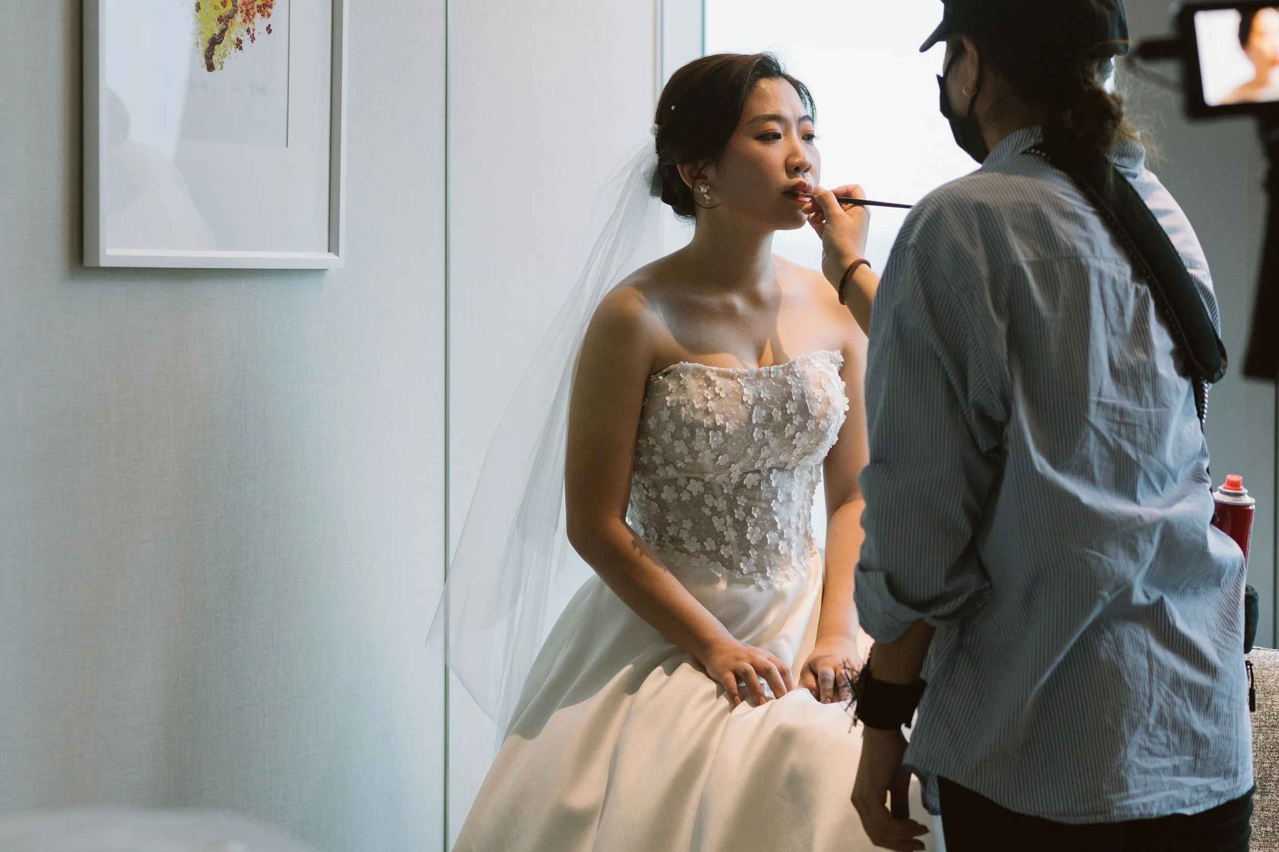 A woman in a wedding dress getting her makeup done by a makeup artist, with a bright window in the background.