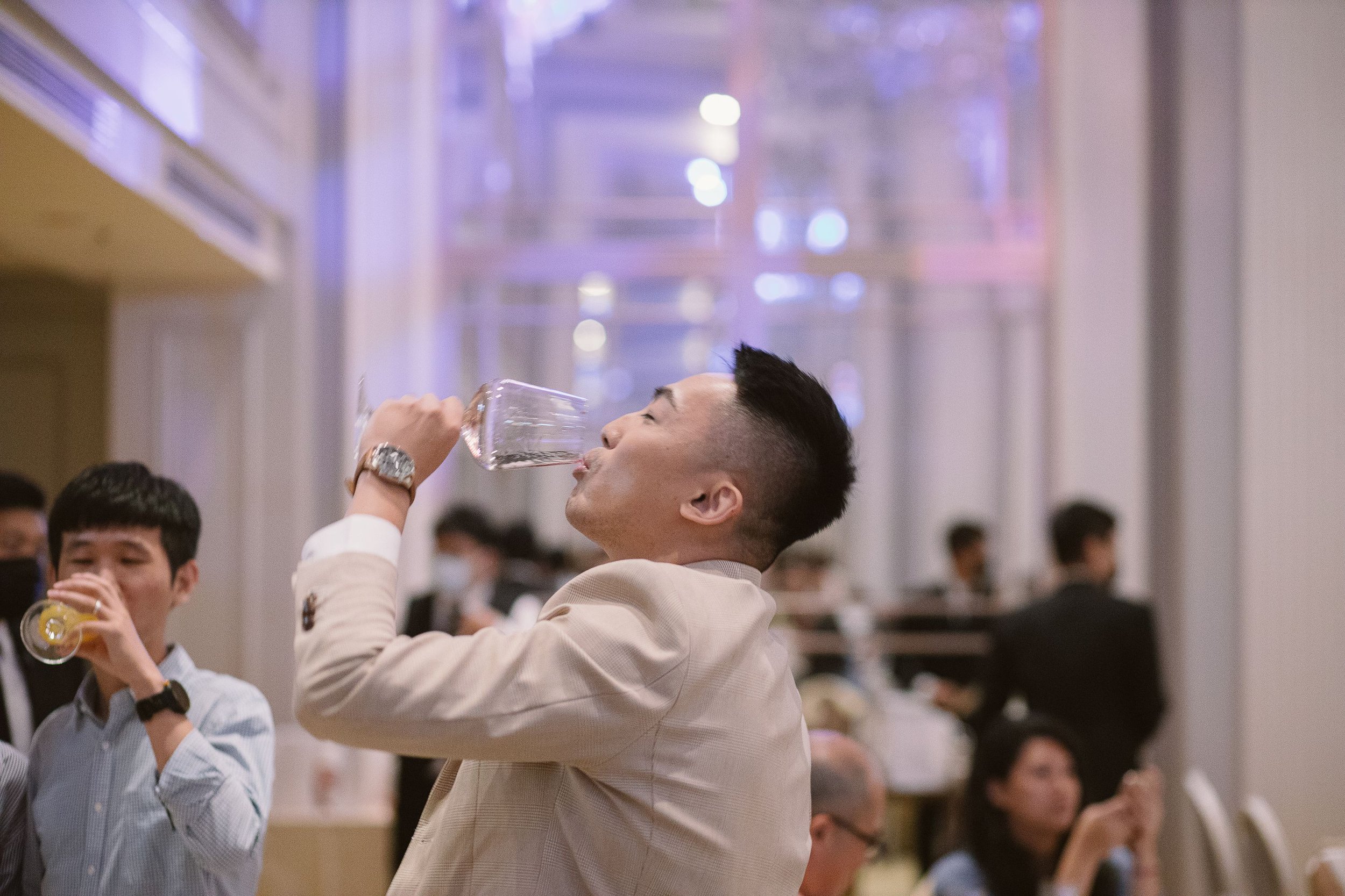 A group of people, mostly men in business attire, are raising wine glasses in a toast at an indoor event with soft lighting and blurred background of other attendees.