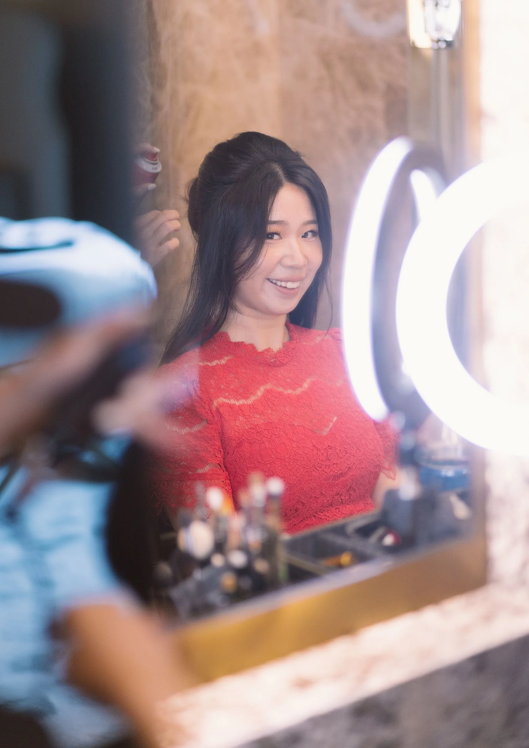 Young woman with long black hair and a big smile, wearing a red lace dress, sitting in front of a ring light while getting her hair styled.