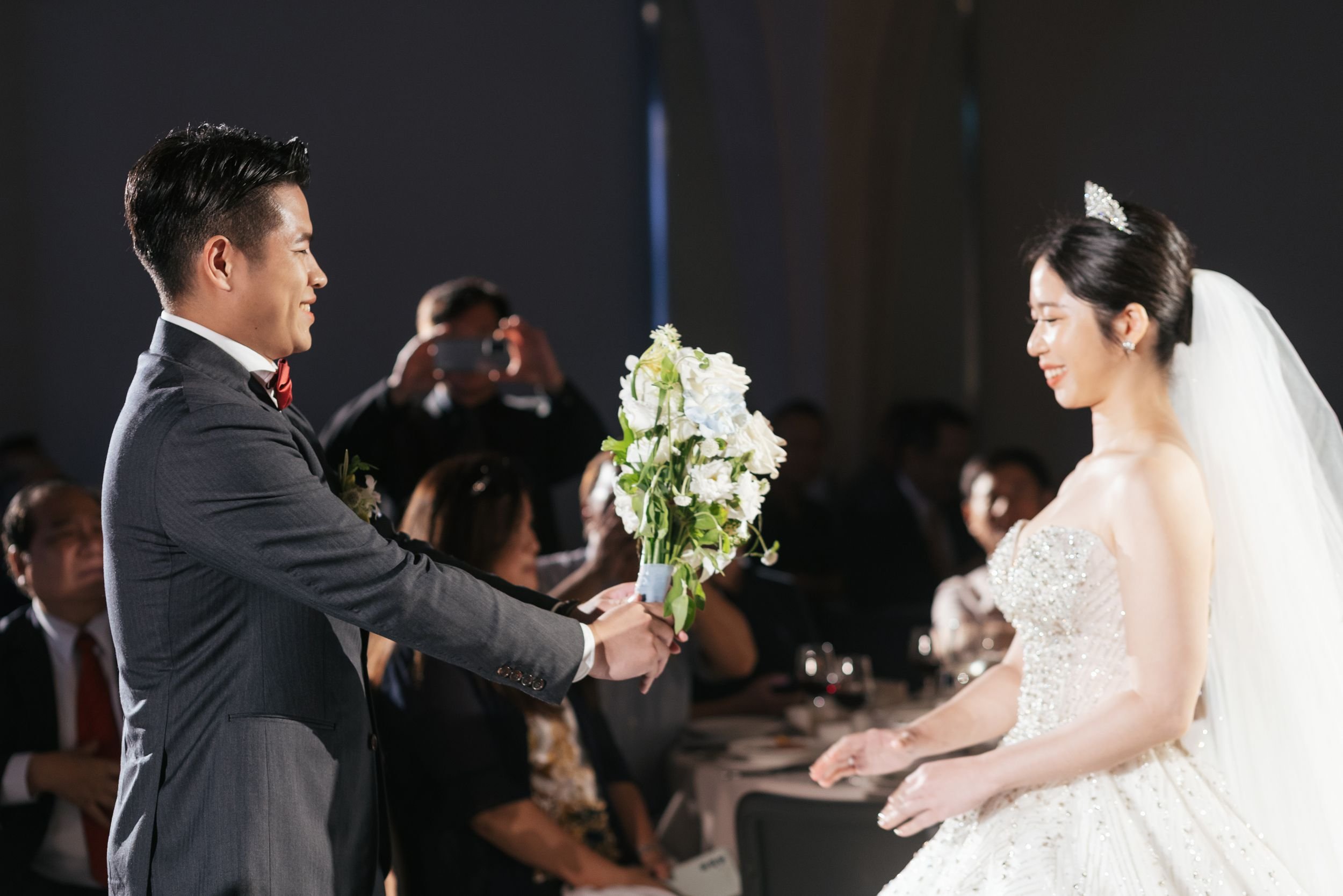 A groom in a tuxedo holding a bouquet of white flowers, smiling at a bride in a wedding dress with a tiara, during a wedding ceremony with guests in the background.