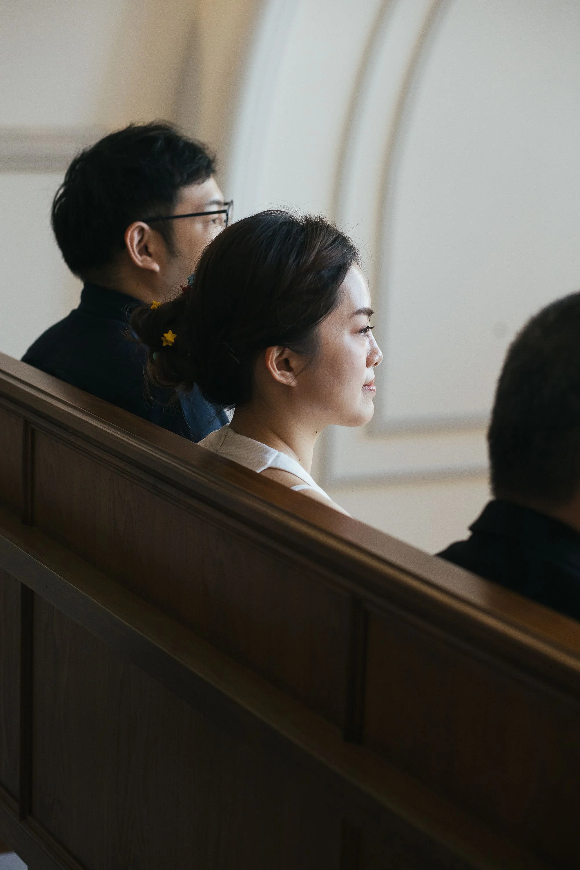 Side view of a woman with dark hair styled in an updo with yellow star-shaped hair accessories, sitting in a church pew next to a man with short dark hair and glasses, both looking forward during a religious service.