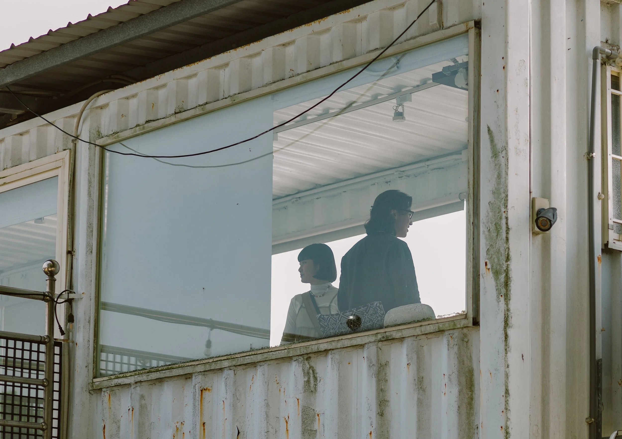 View through a building window showing two people, one with glasses and a woman, standing inside a structure with moss and rust on the exterior walls and some electrical wiring.