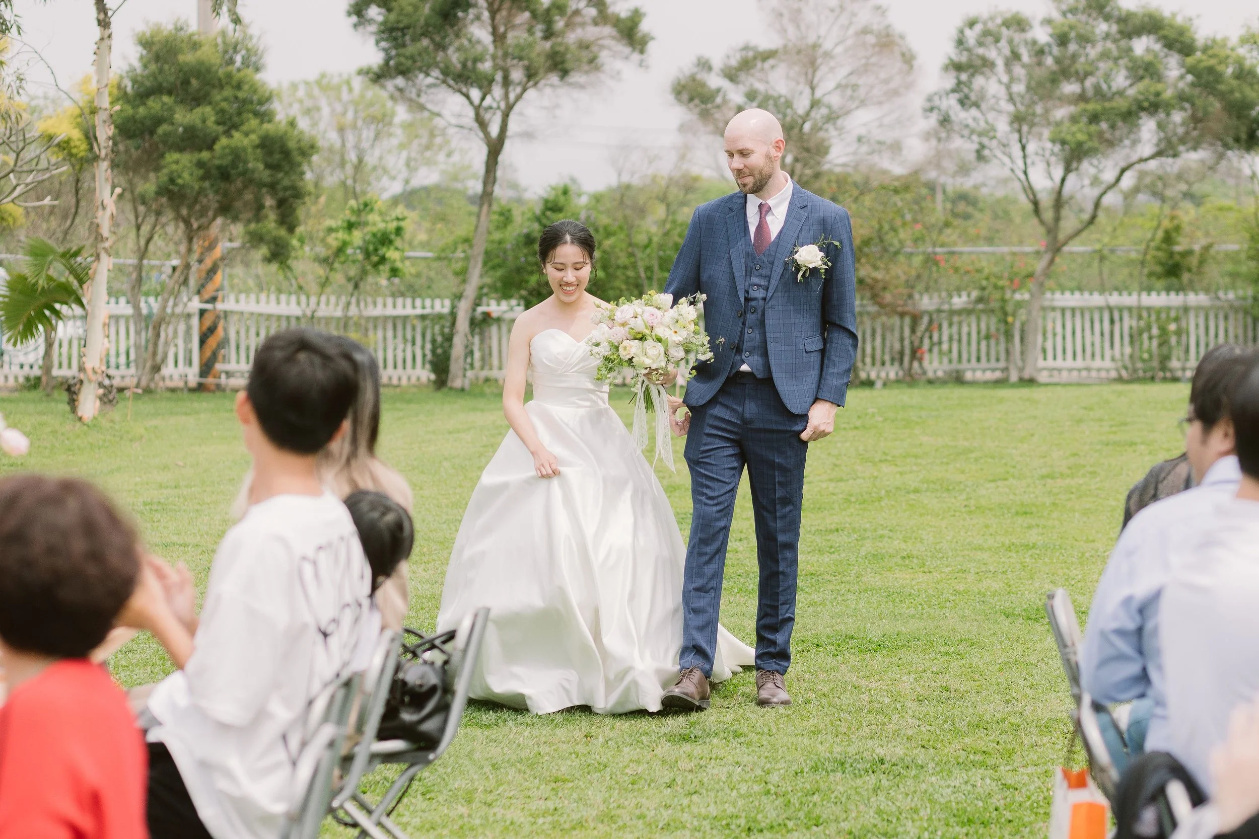 A bride and groom stand outdoors during their wedding ceremony, surrounded by seated guests. The bride holds a bouquet of flowers, and the groom wears a blue suit with a white flower boutonniere. The background shows a white picket fence and trees.