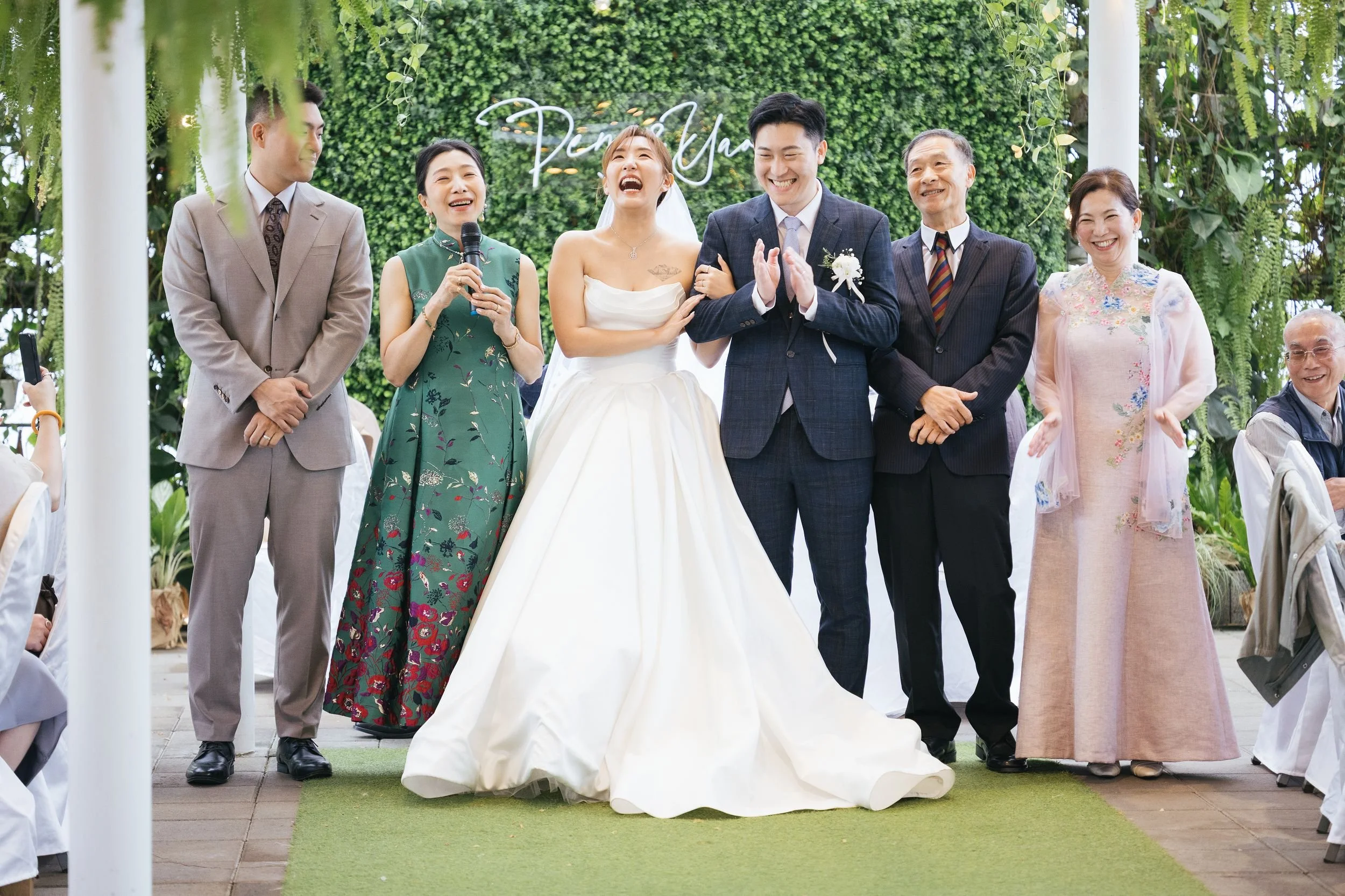 A group of people at a wedding reception, with a bride in a white wedding dress and a groom in a gray suit, surrounded by family and friends, smiling and laughing in a decorated outdoor setting with greenery and hanging plants.