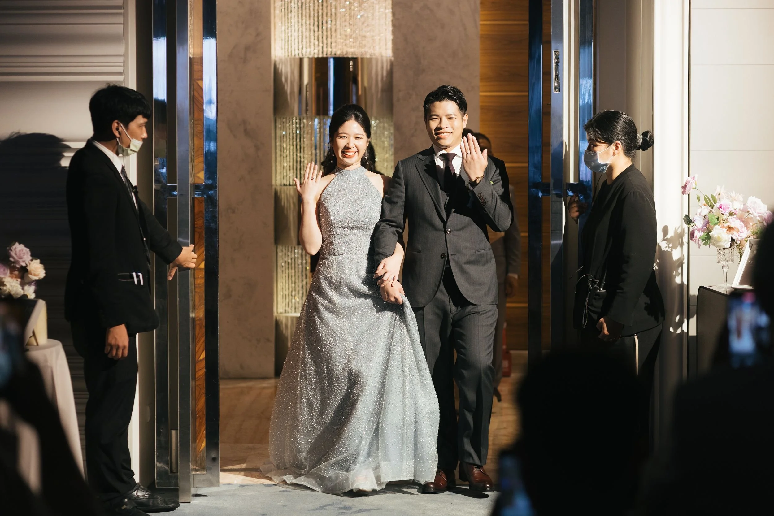 A couple, dressed in formal attire, smiling and holding hands as they walk through a doorway at a wedding reception, with event staff on either side.