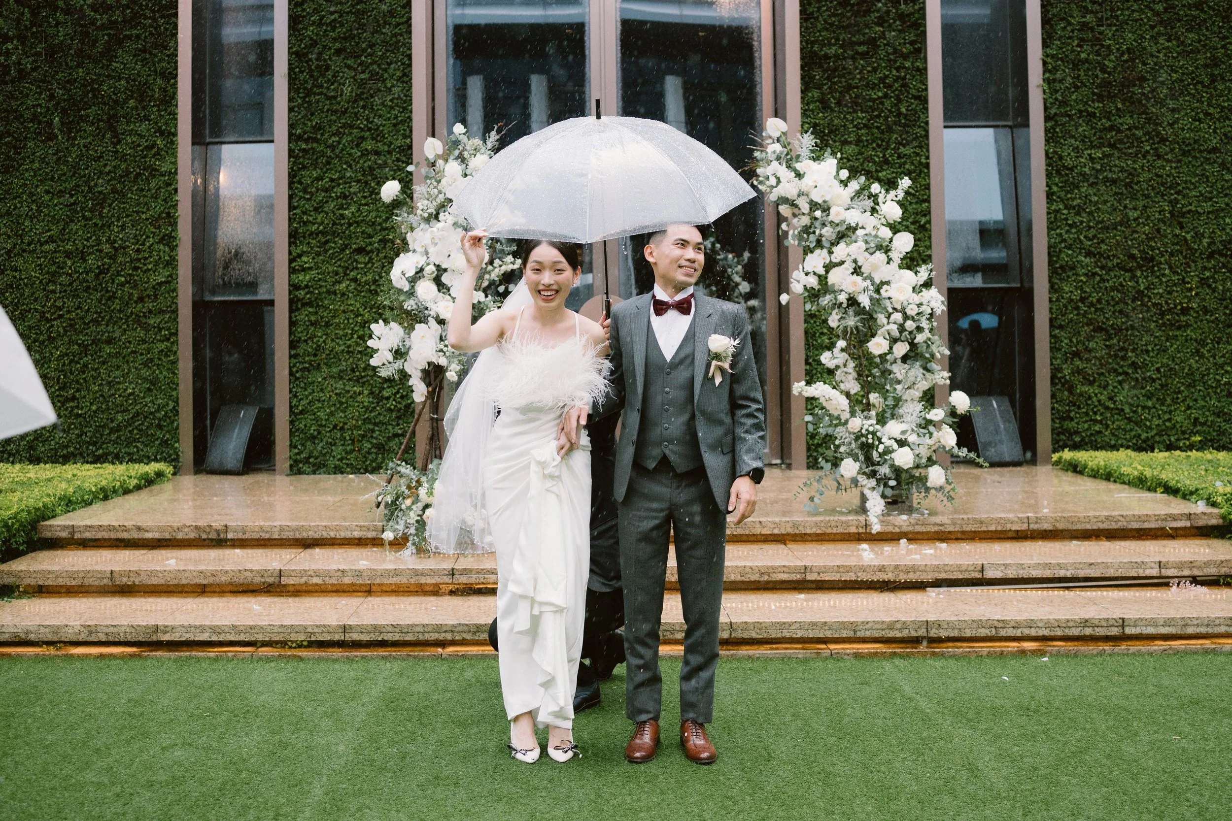 A newlywed couple standing on stone steps outdoors during a rainy wedding, holding a transparent umbrella. The bride is wearing a white wedding dress and the groom is wearing a gray suit with a burgundy bow tie. They are smiling and a floral arch wit