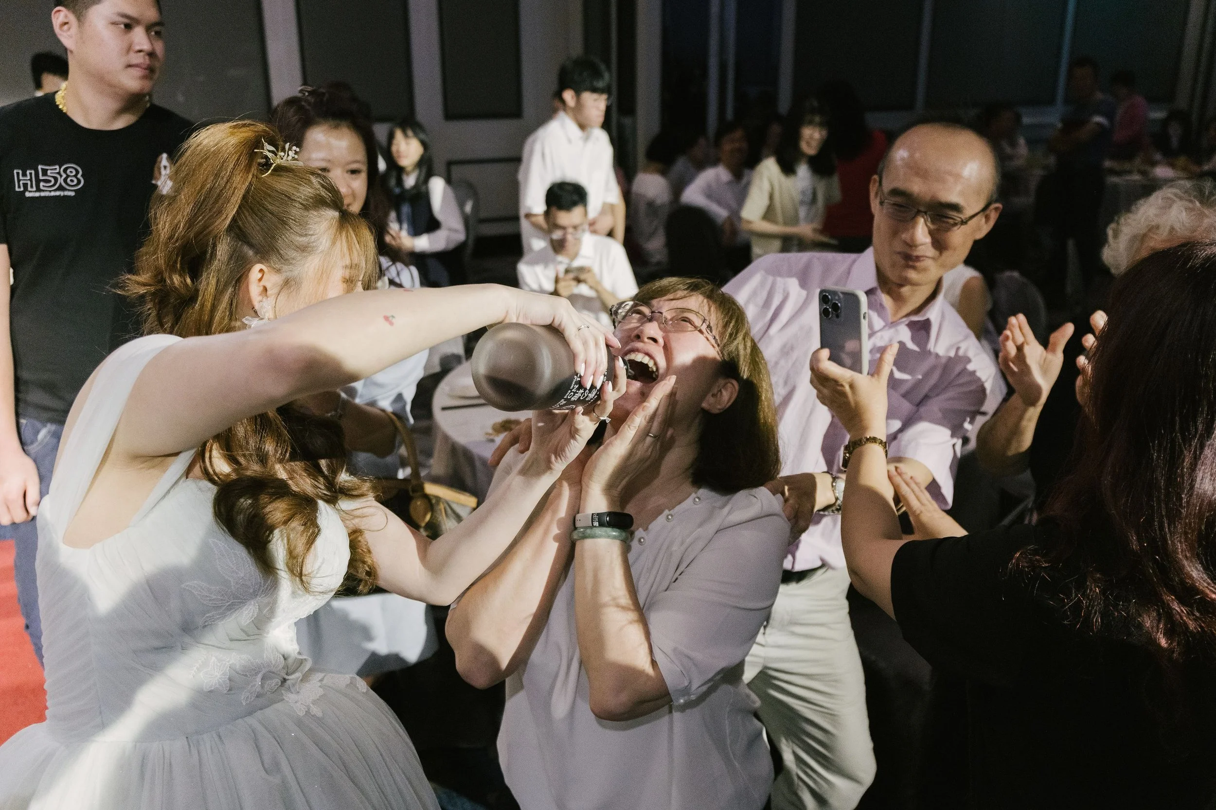 A group of people gathered around at a social event, with a woman drinking from a bottle, smiling, as others watch and take photos in a dimly lit room.