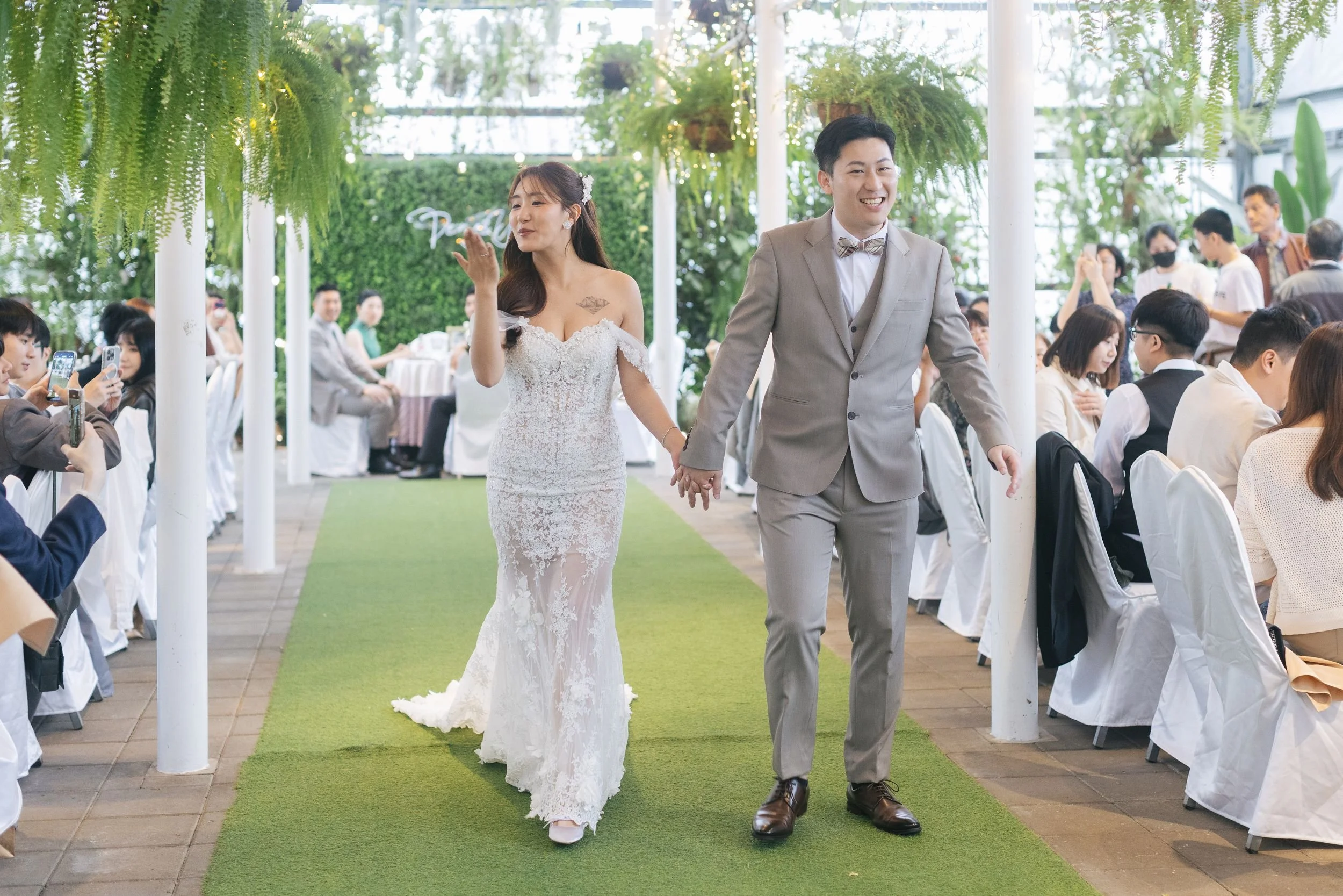 A bride and groom walking hand in hand down an aisle at their wedding reception, surrounded by seated guests in a bright, green indoor venue with hanging plants.