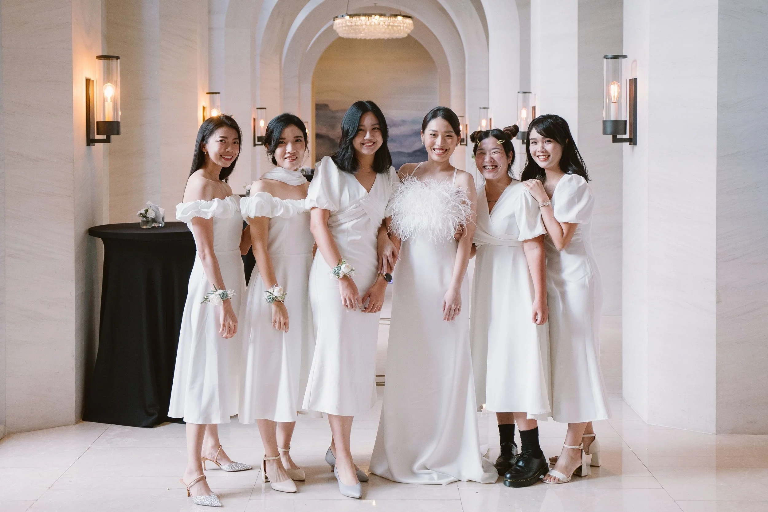 Group of six women dressed in white dresses, standing in a hallway with elegant lighting and marble walls, smiling for the camera.