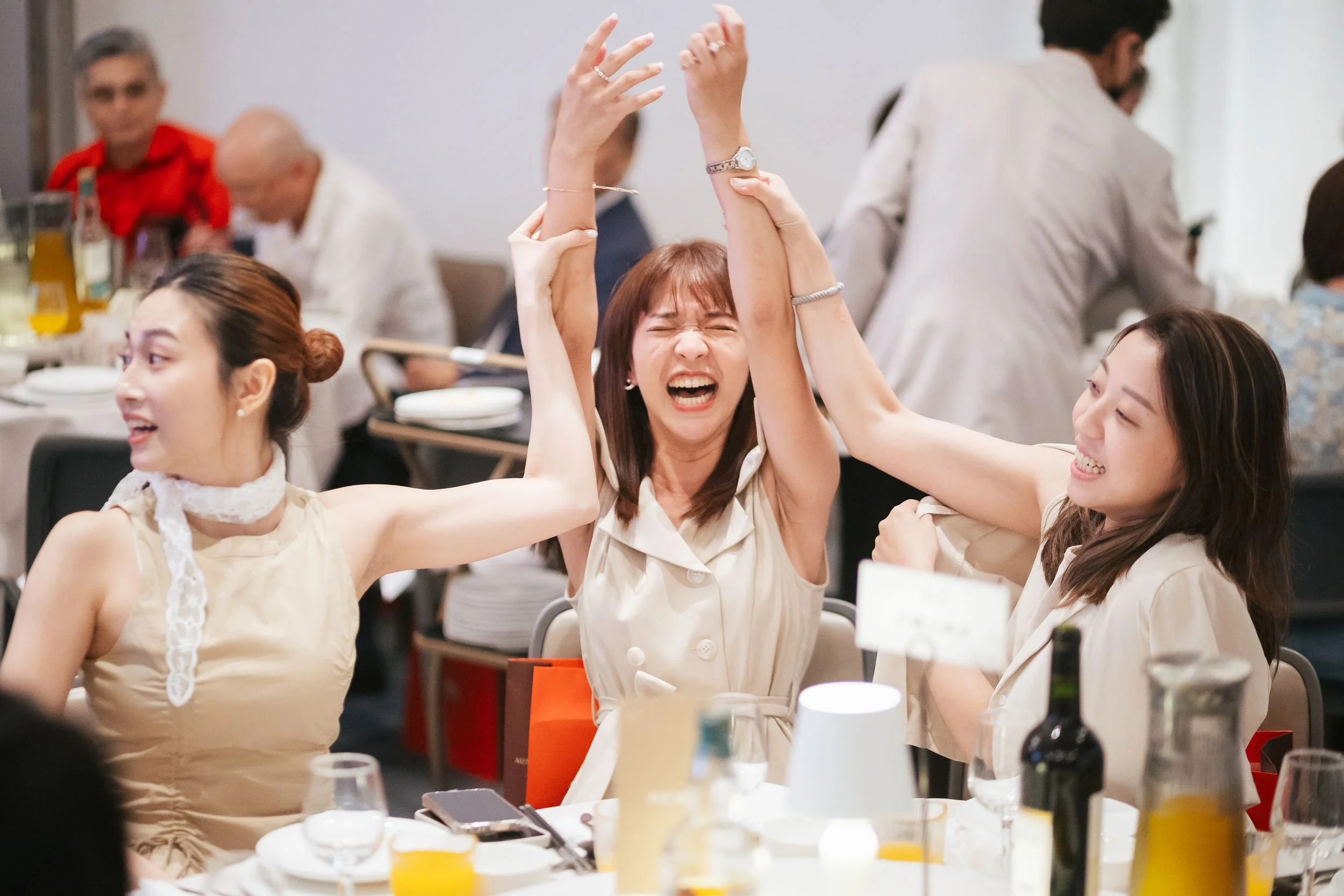 Three women are enjoying themselves at a dining table, with the woman in the middle raising her hands joyfully and laughing. The women are dressed in light-colored outfits, and the table is set with drinks, bottles, and utensils, indicating a celebra