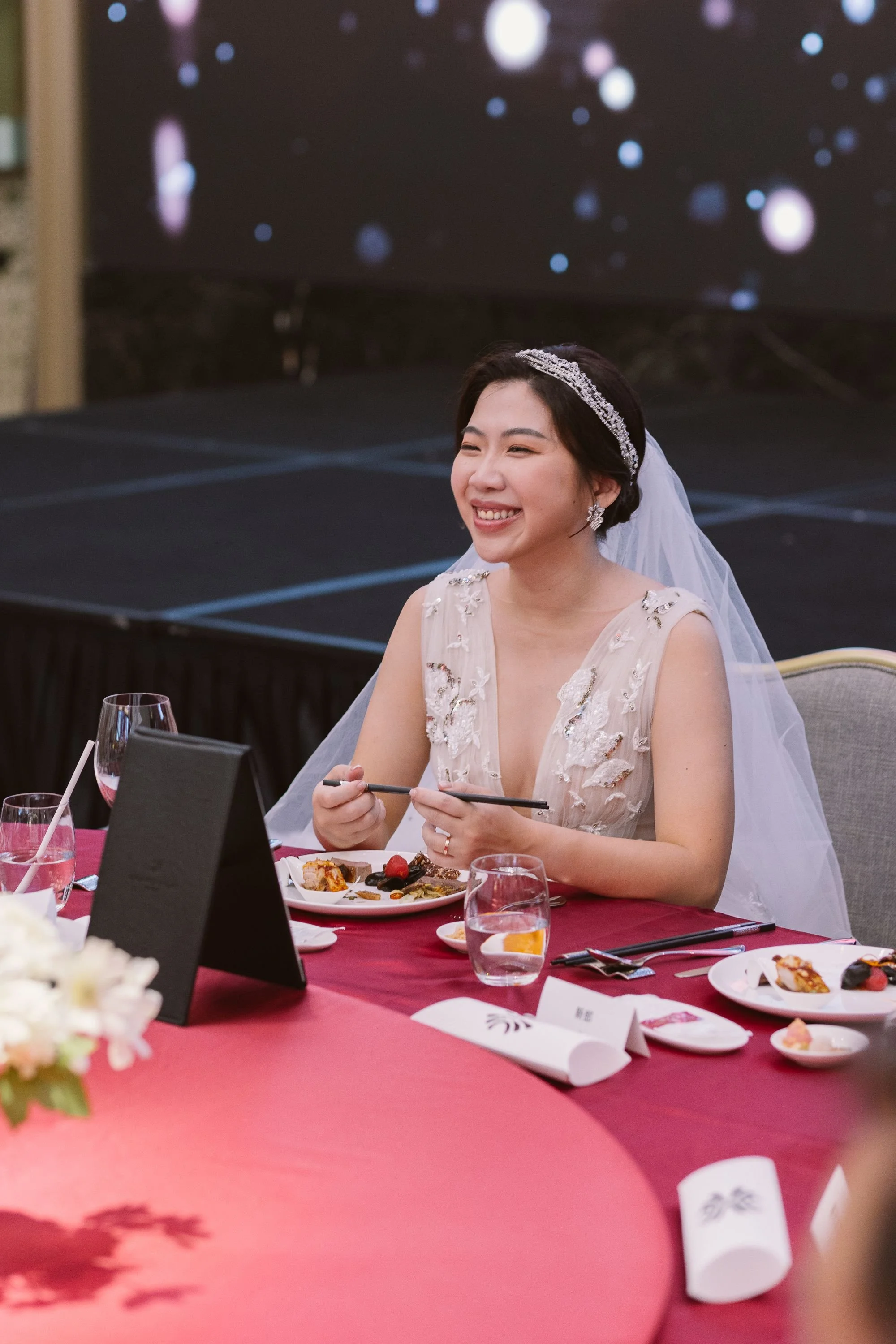 A woman in a wedding dress with a tiara and veil smiles while sitting at a table set for a celebration, with food and drinks in front of her.
