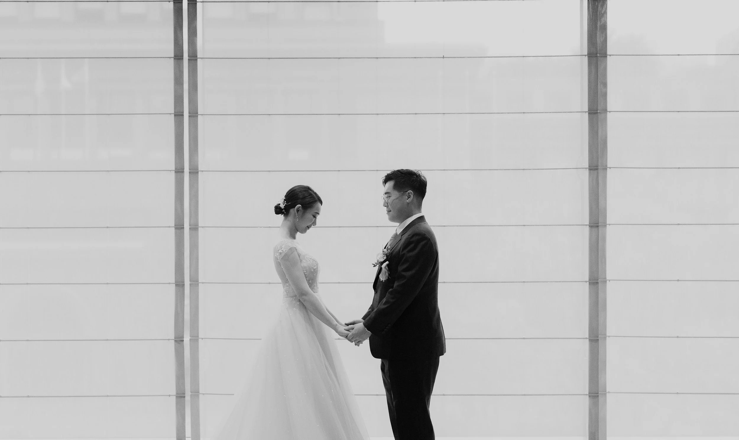 A black and white photo of a bride and groom holding hands and smiling at each other, standing in front of a modern, minimalistic wall with horizontal lines.