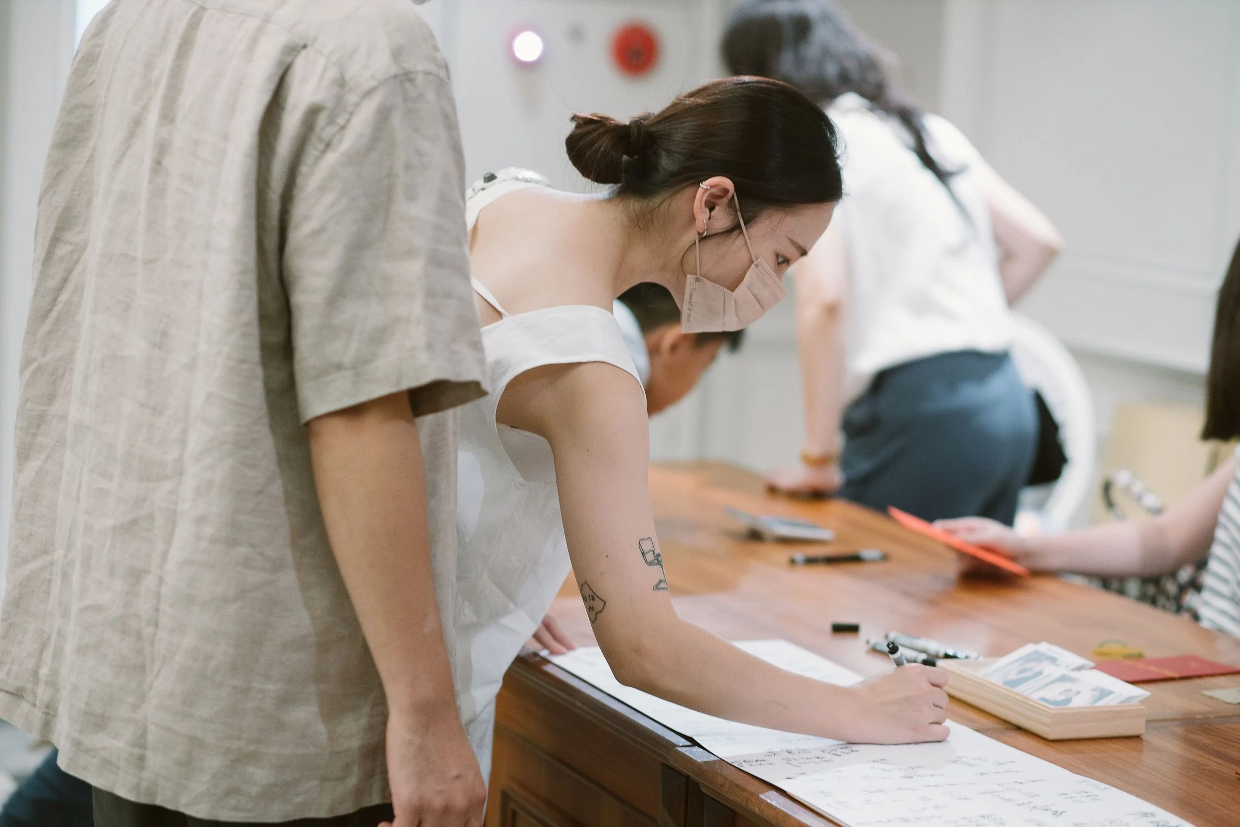 People wearing face masks and leaning over a wooden table, working with papers and writing materials in a bright room.