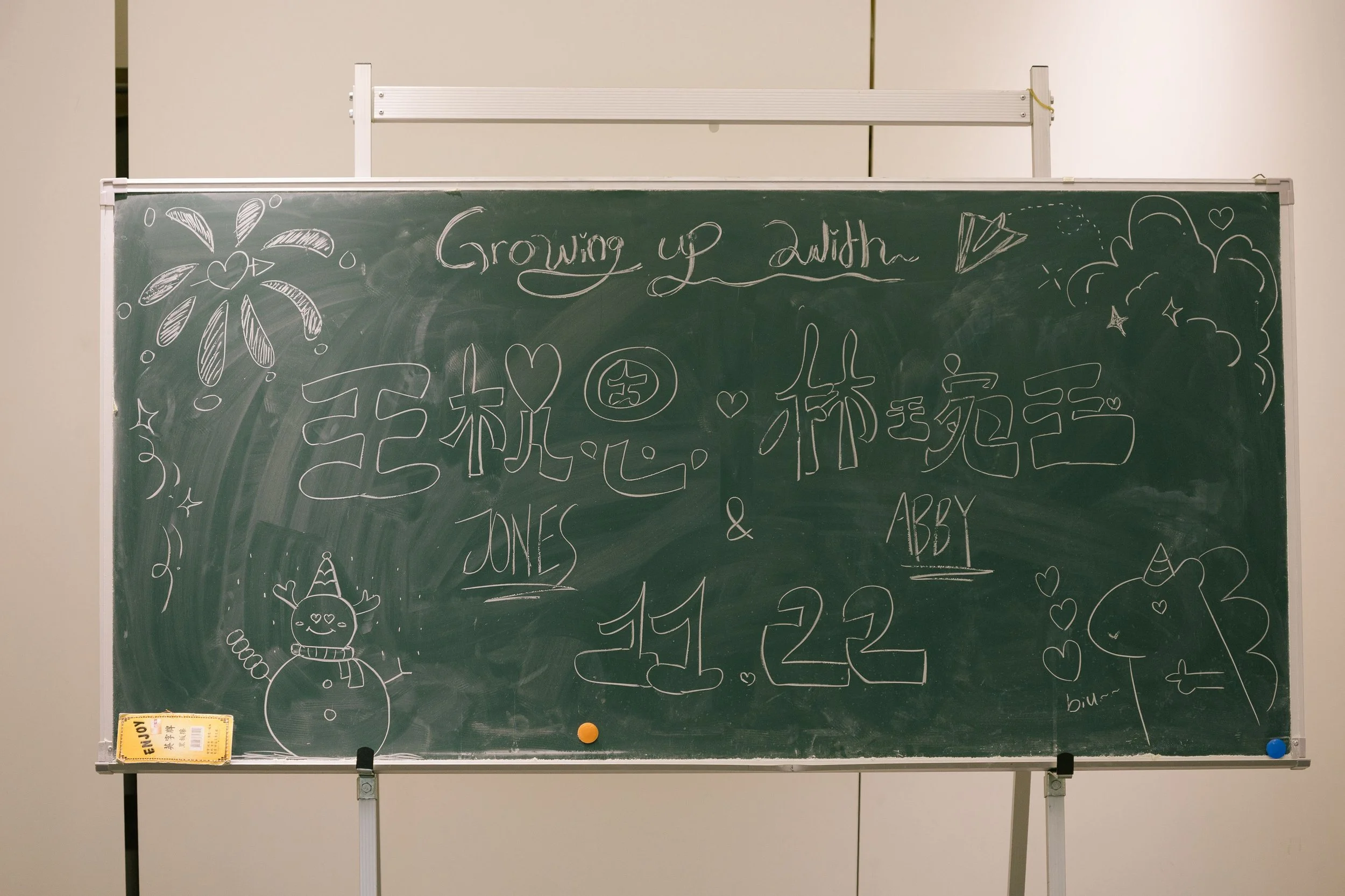 A green chalkboard with decorative drawings including a snowman, a reindeer, a paper airplane, and various hearts and stars. The words "Growing up with" are written at the top, and there are some names and numbers below.