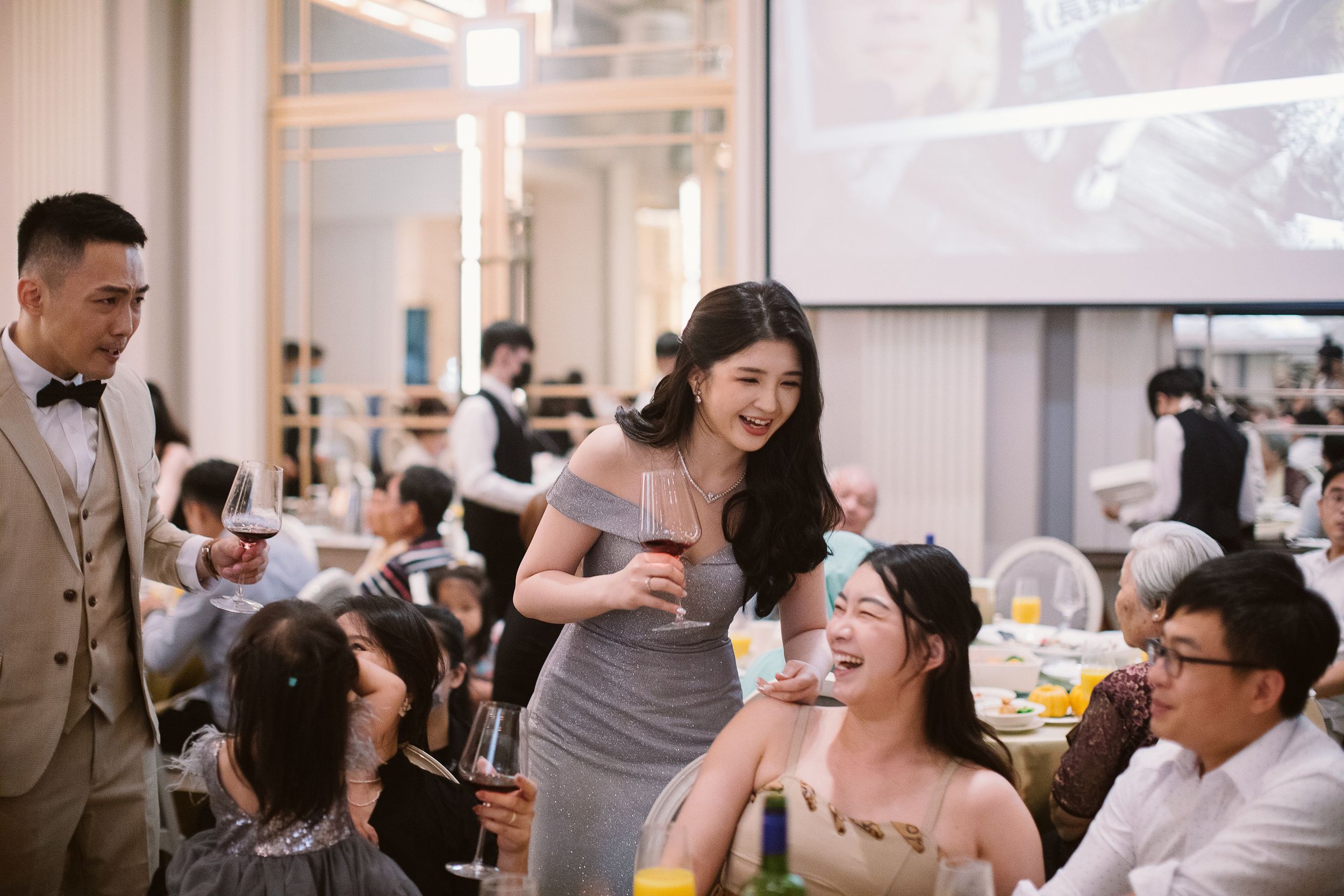 Guests at a formal event, with a woman in a gray dress smiling and holding a glass of red wine, and a man in a beige suit with a bow tie also holding a glass of red wine, in a decorated banquet hall with waitstaff in black vests and white shirts serv