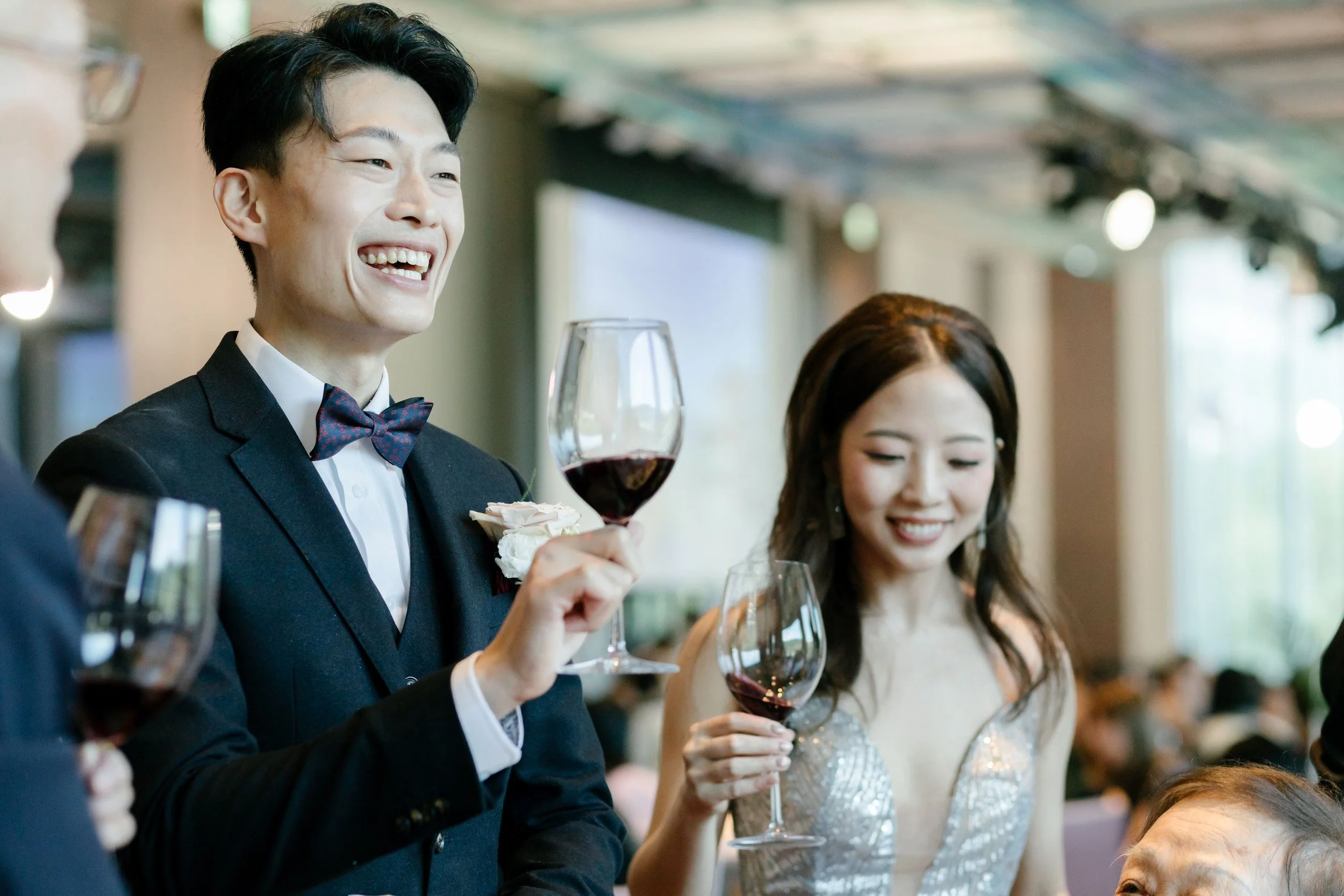 A smiling man in a tuxedo with a bow tie holding a glass of red wine at a formal event, with women in elegant dresses around him.