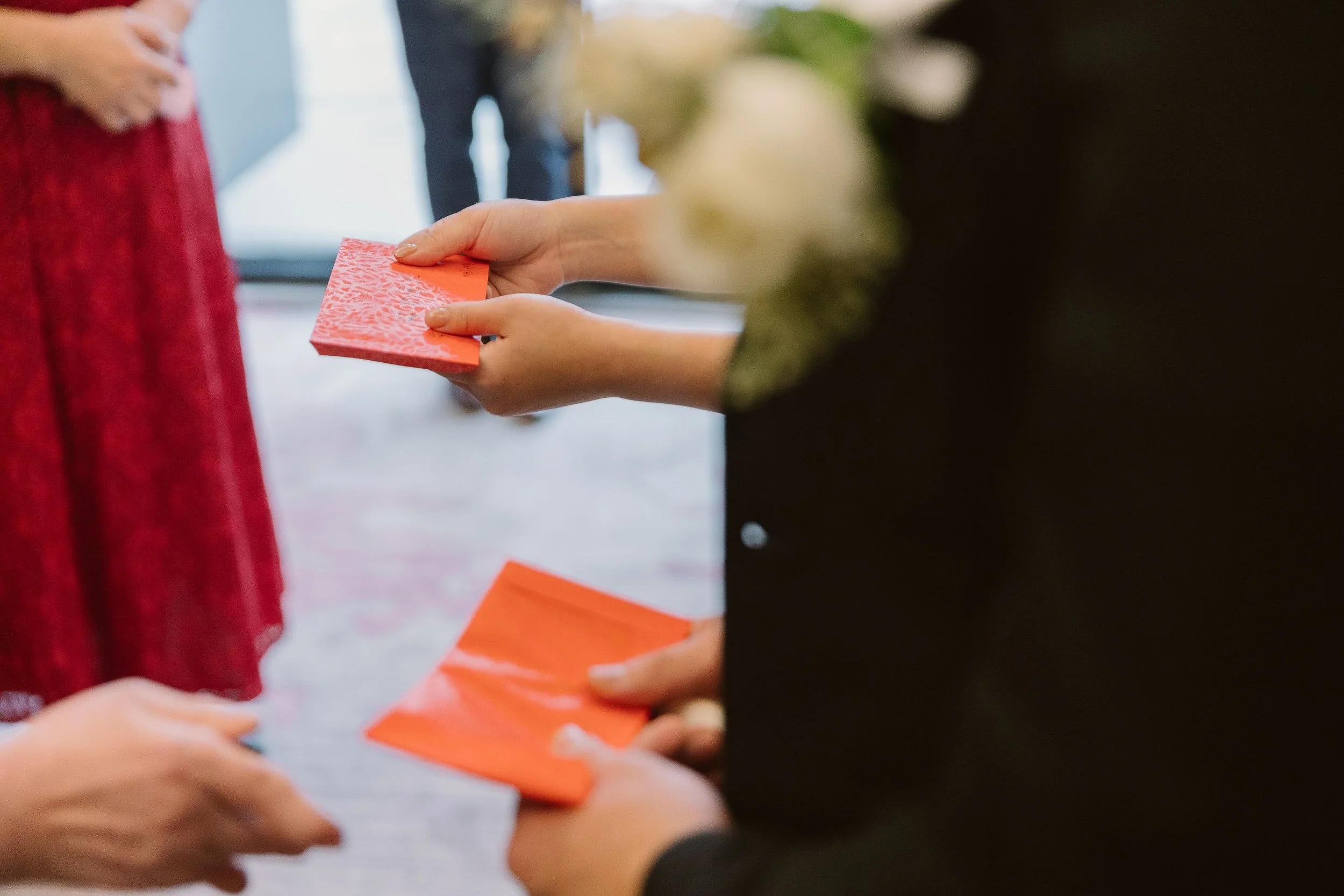 Close-up of a person’s hands exchanging or holding small orange and pink rectangular cards or paper snippets, with other people in the background.