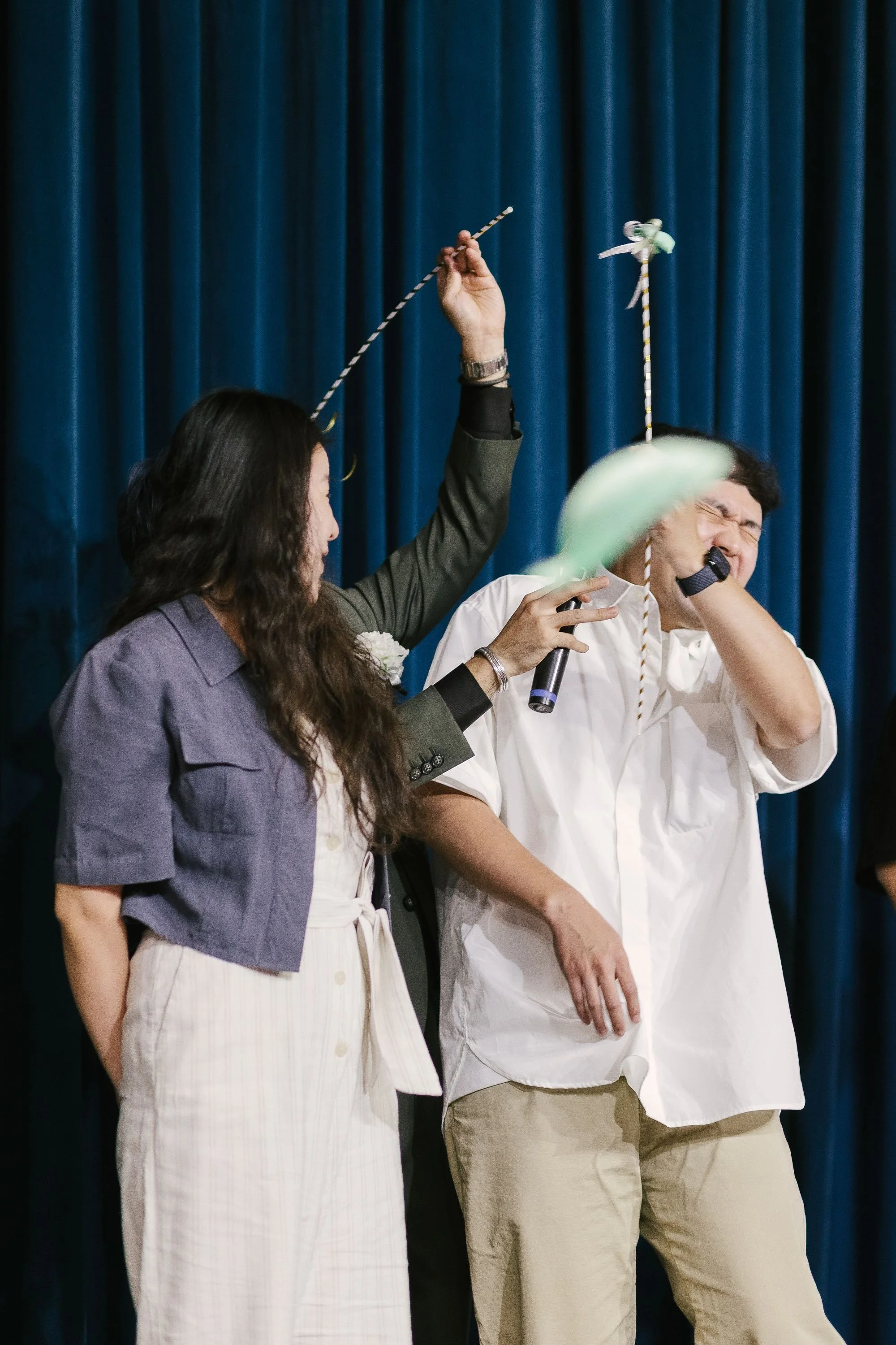 Two people appear to be participating in a game or activity involving spinning or balancing objects on sticks, with a blue curtain in the background.