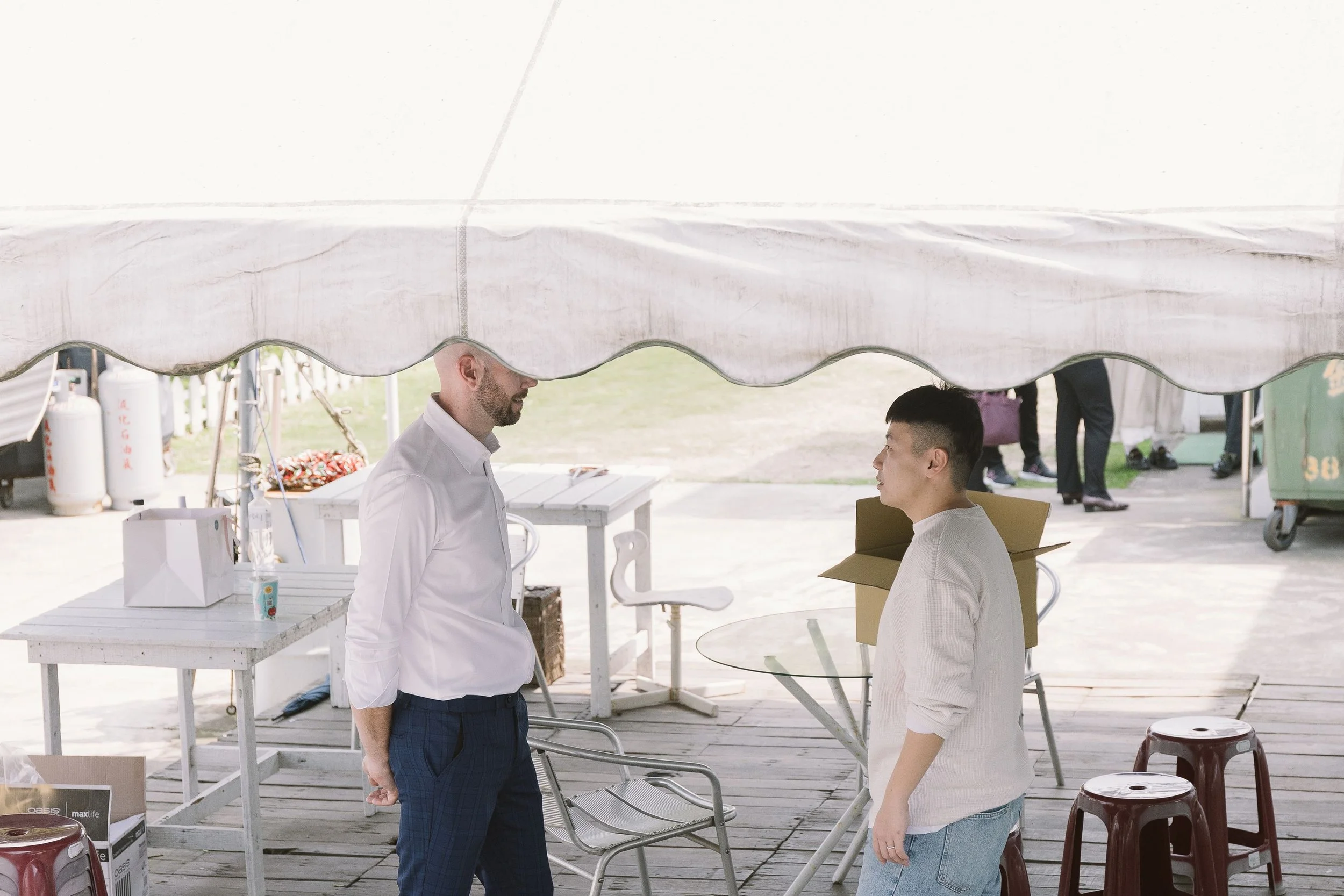 Two men having a conversation under a white canopy outdoors, with chairs, tables, and supplies around them.