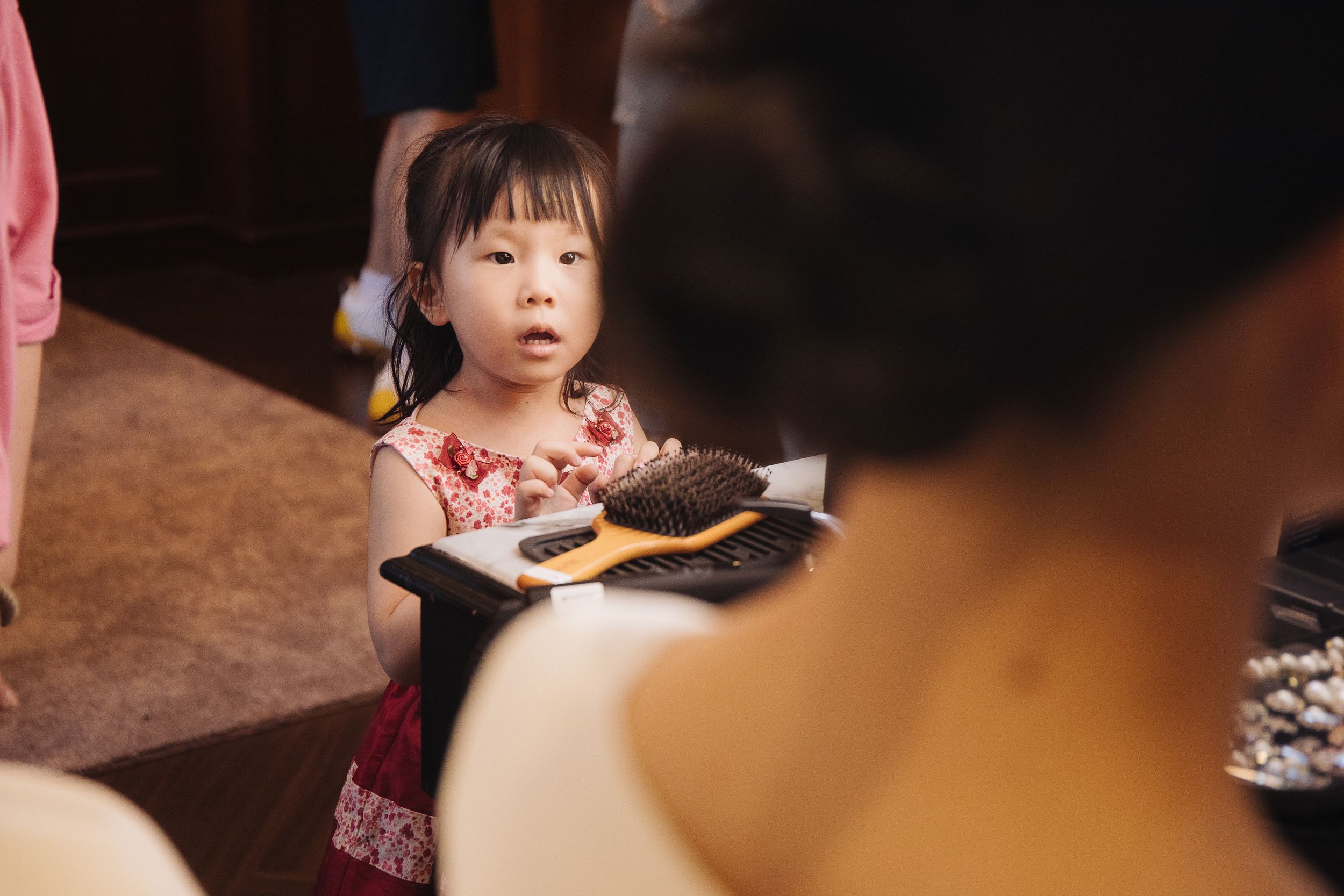 A young Asian girl with dark hair and bangs wearing a pink floral dress with bows, sitting at a table with a hairbrush and a hair dye palette in front of her. She appears surprised or curious, engaging with someone or something off-camera.