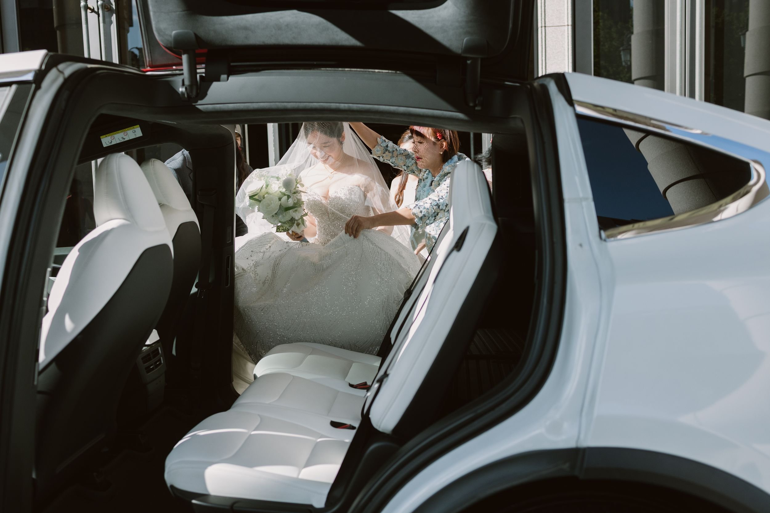 A bride in a wedding dress and veil is sitting in the back seat of a car, smiling as a woman adjusts her veil from outside the car. Another person partially visible is in the background.