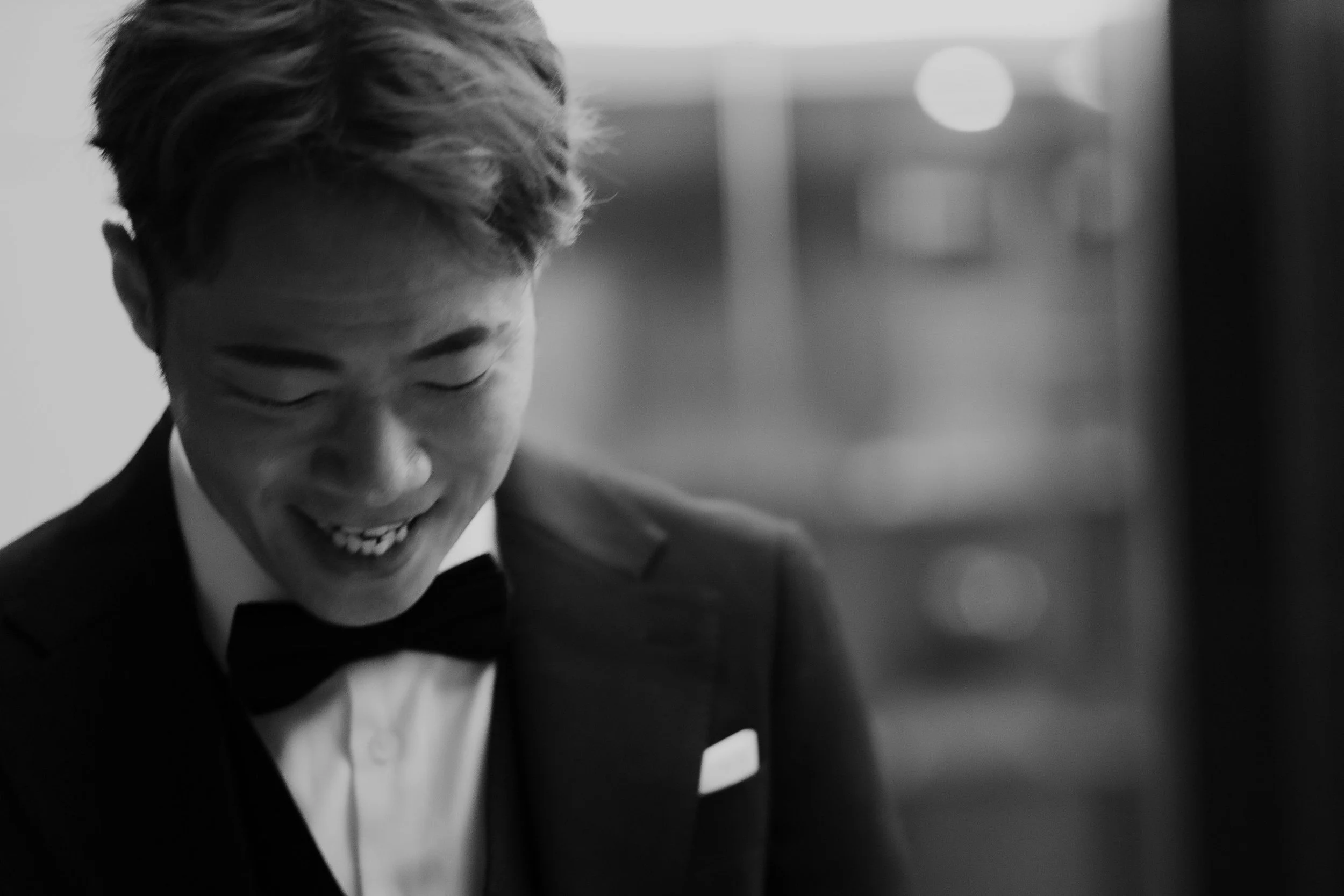 Black and white photo of a person in a tuxedo, smiling with eyes closed, with a blurred background.
