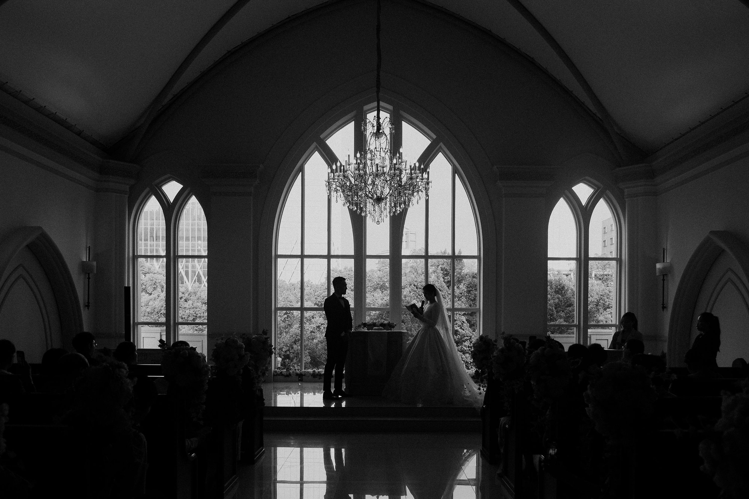 Silhouetted couple standing before an officiant during a wedding ceremony in a church with large arched windows and a chandelier.