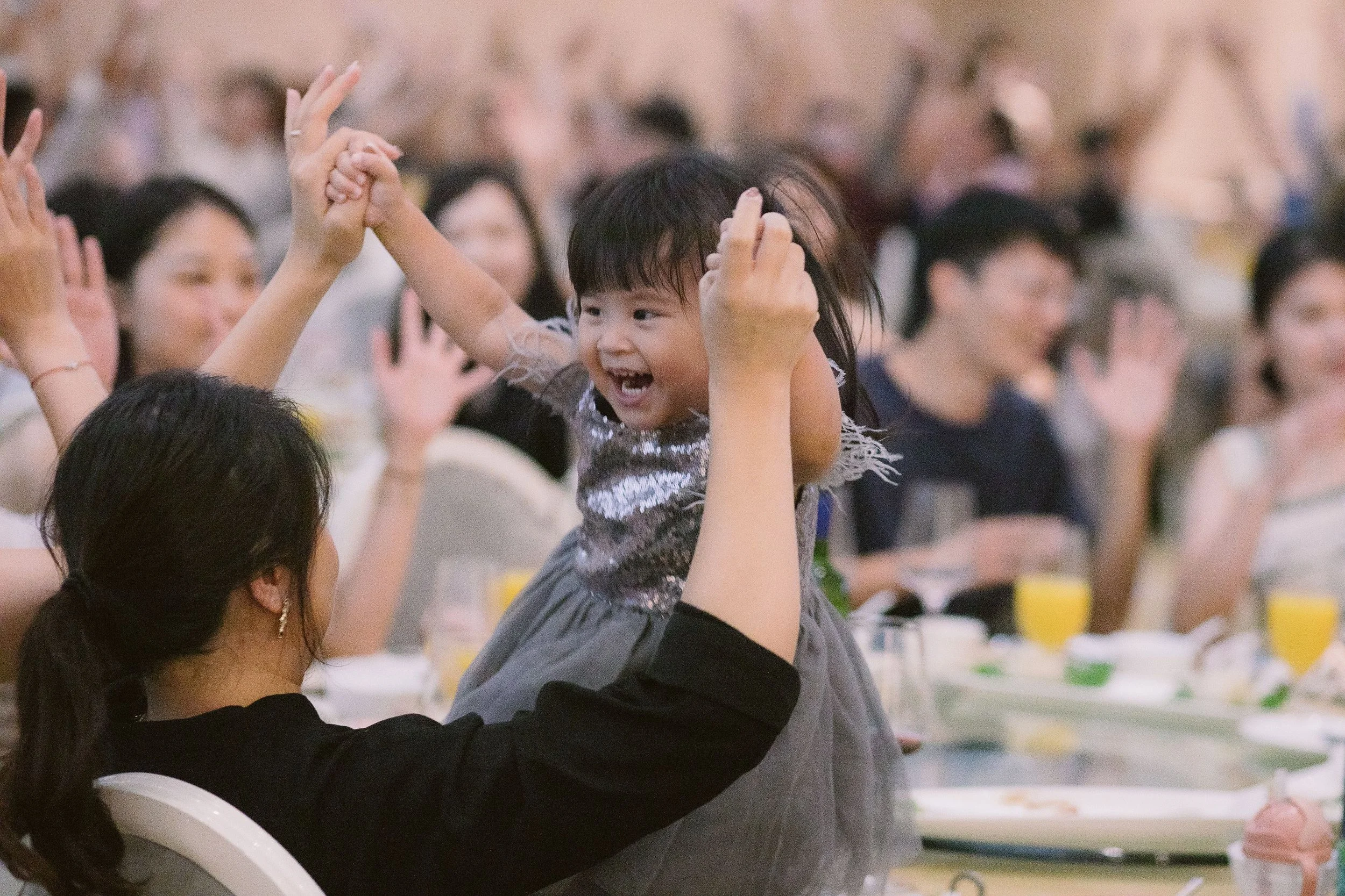 A young girl with dark hair and bangs playing at a table during a gathering, smiling with her hands raised, surrounded by an audience.