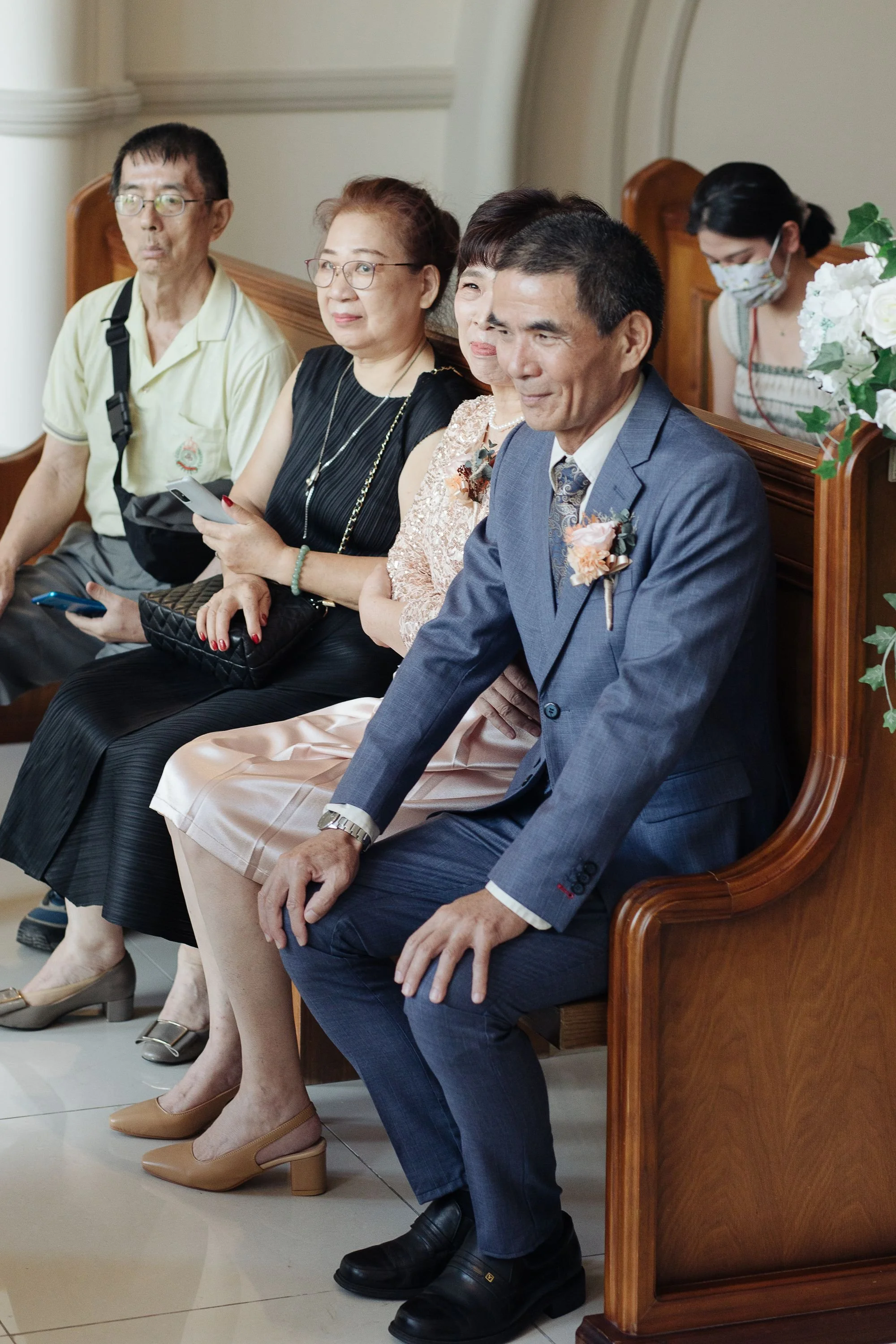 A group of five people sitting in a row, dressed formally, inside a church or similar setting, with some holding phones. The man in the blue suit has a boutonniere, indicating a wedding or special occasion.