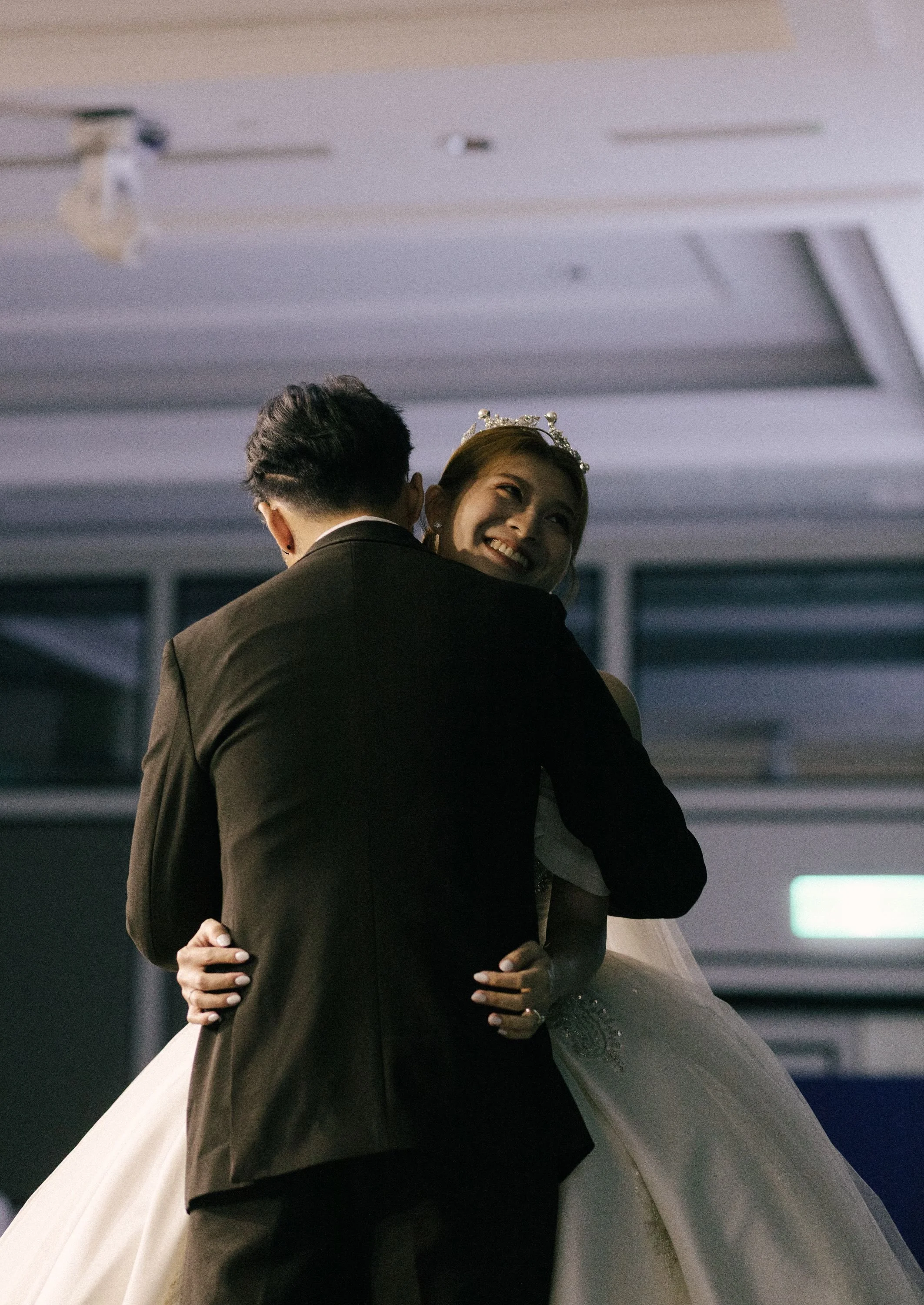 A bride and groom dancing, with the bride wearing a white wedding gown and a tiara, and the groom in a dark suit, in an indoor setting.