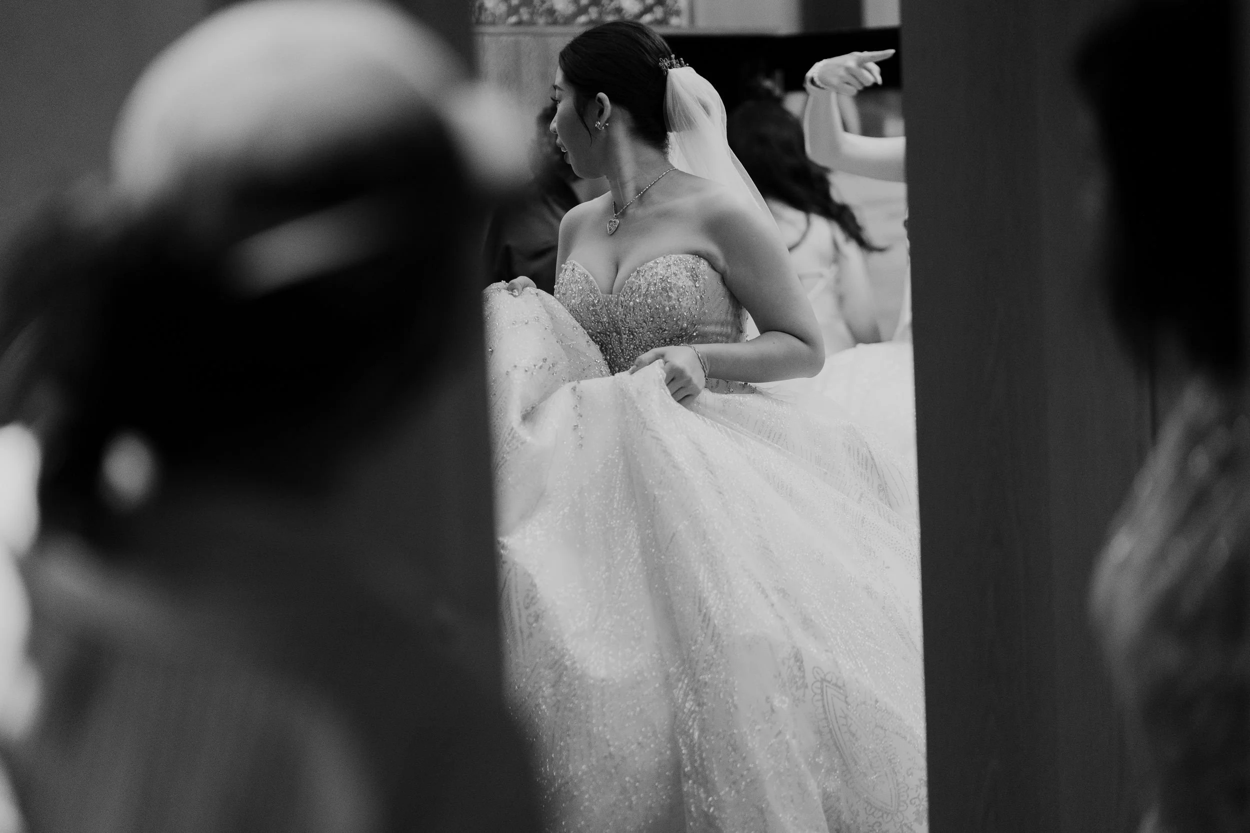 A black and white photo of a bride in a strapless wedding gown with beaded details, sitting and surrounded by guests at a wedding reception. She is wearing a veil and a necklace.