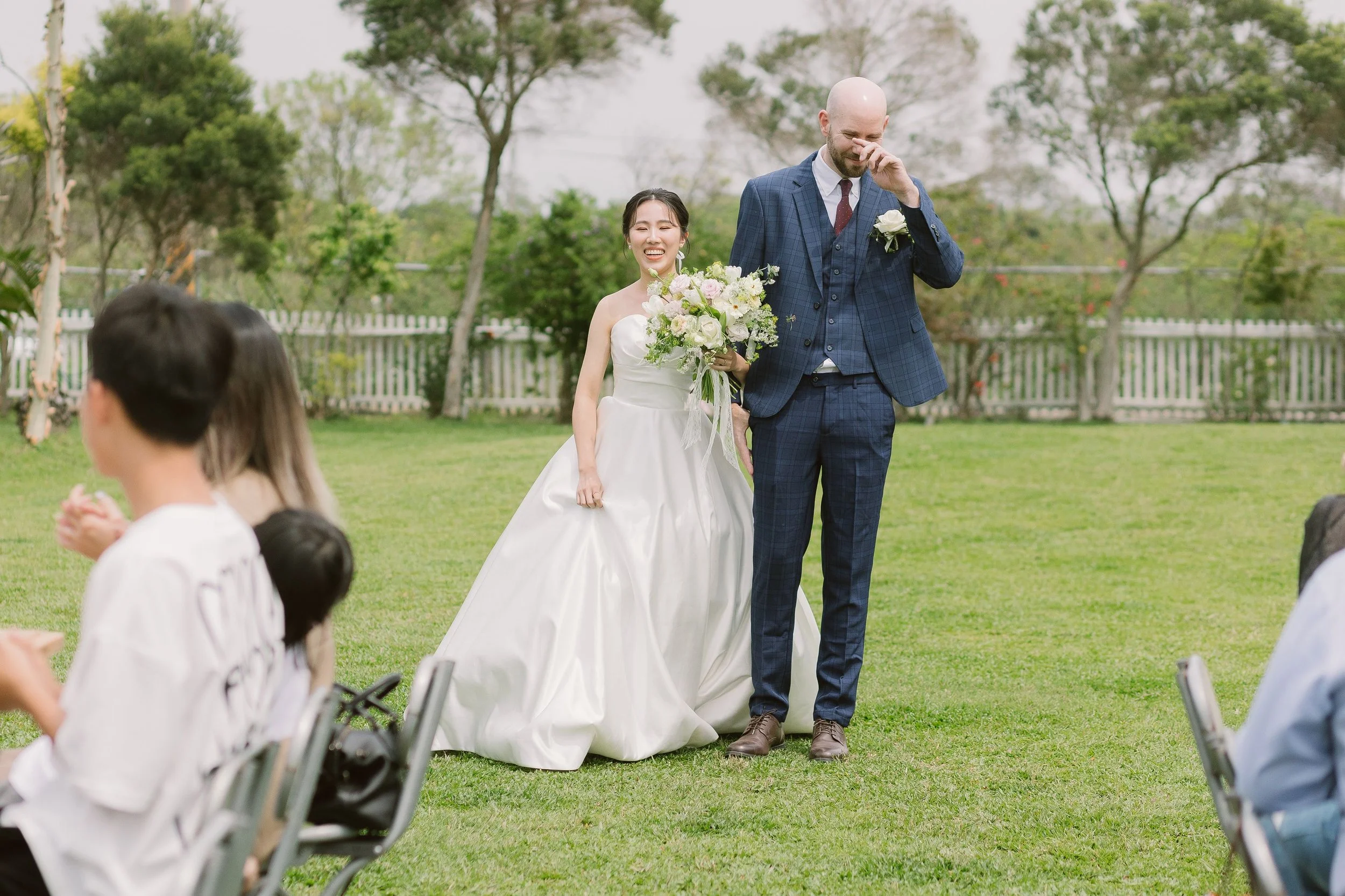 A bride in a white wedding dress holding a bouquet of flowers, smiling, walking arm-in-arm with a bald groom in a blue checkered suit, at an outdoor wedding ceremony.