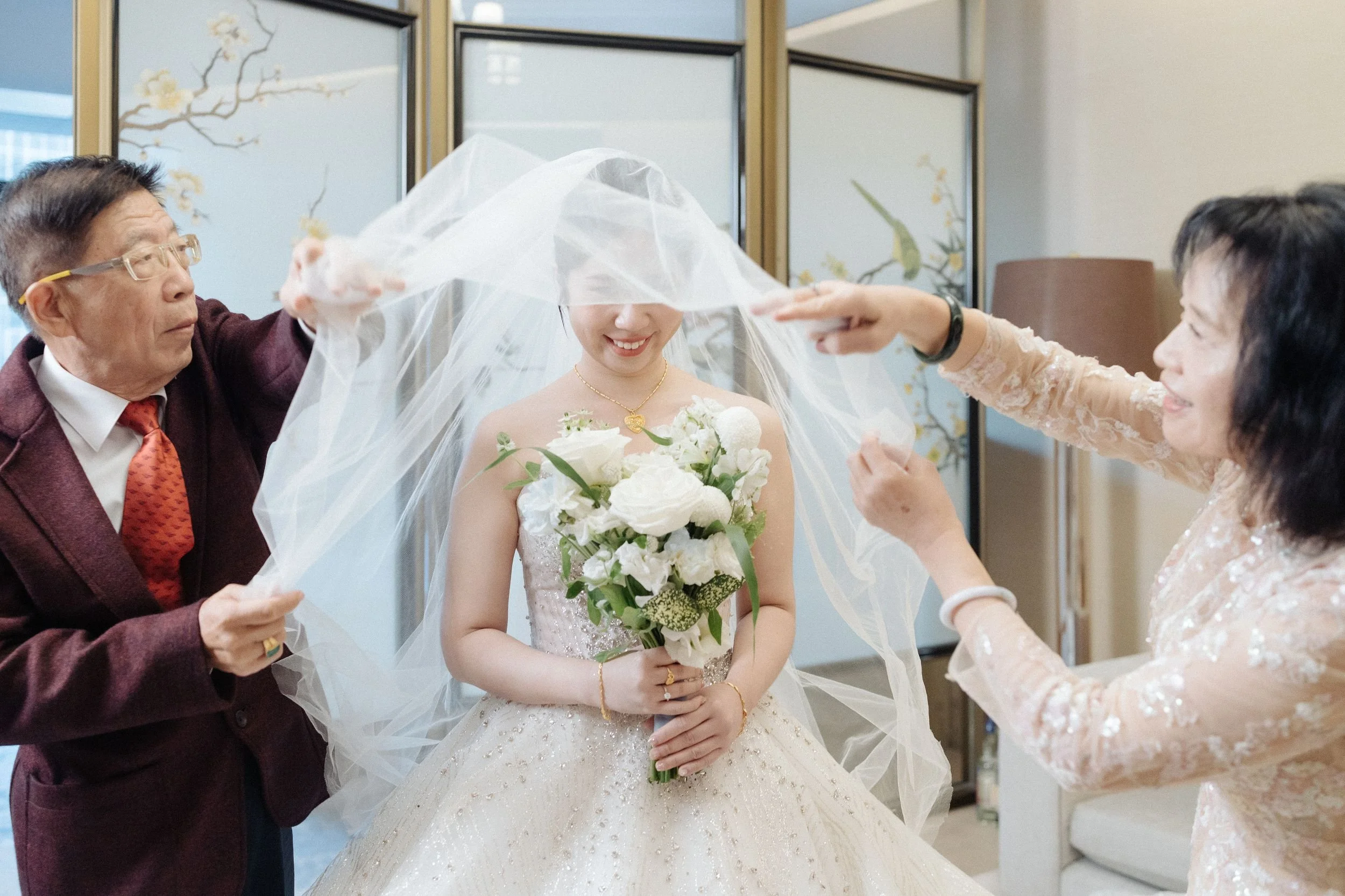 A bride in a wedding dress holding a bouquet of white flowers is being assisted by two women, while an older man helps with her veil, in an indoor setting.