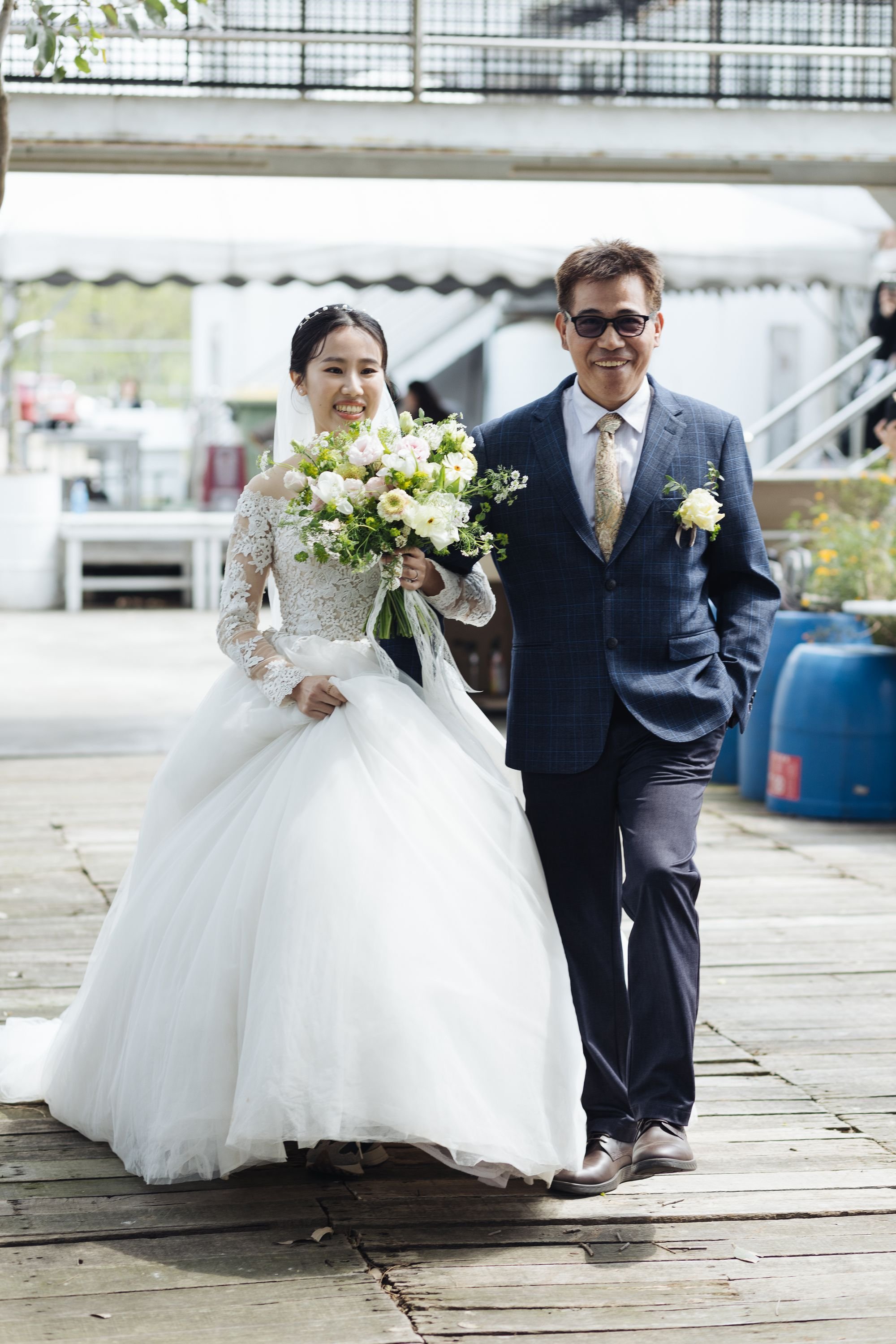 A bride and a man, possibly her father, walking together outdoors on a wedding day. The bride is dressed in a white wedding gown and is holding a bouquet of flowers. The man is dressed in a dark plaid suit with sunglasses.