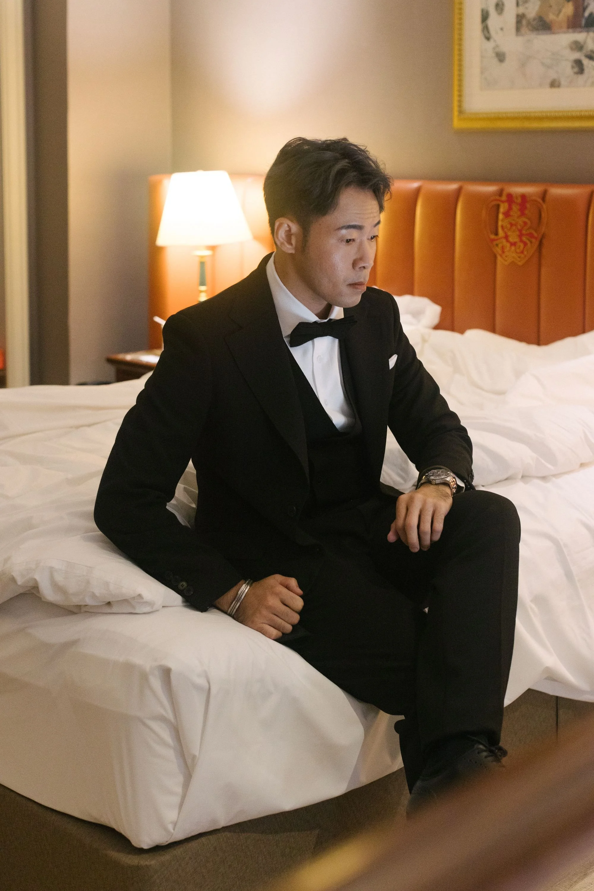 A man in a tuxedo sitting on a bed in a hotel room with a serious expression.