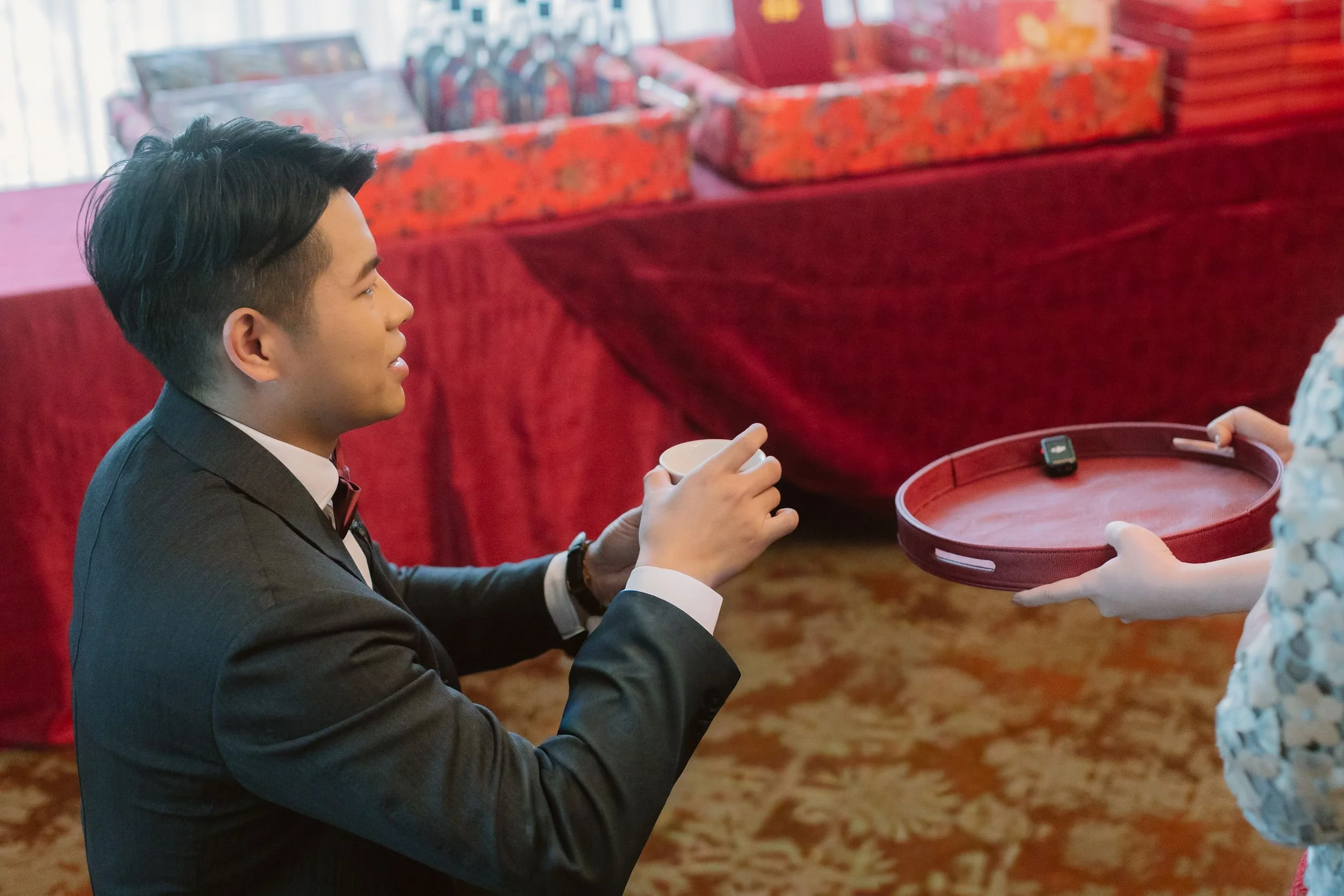 A man in a tuxedo with a bow tie is sitting at a table, holding a small cup with both hands, as someone off-camera hands him a tray with a remote control on it.
