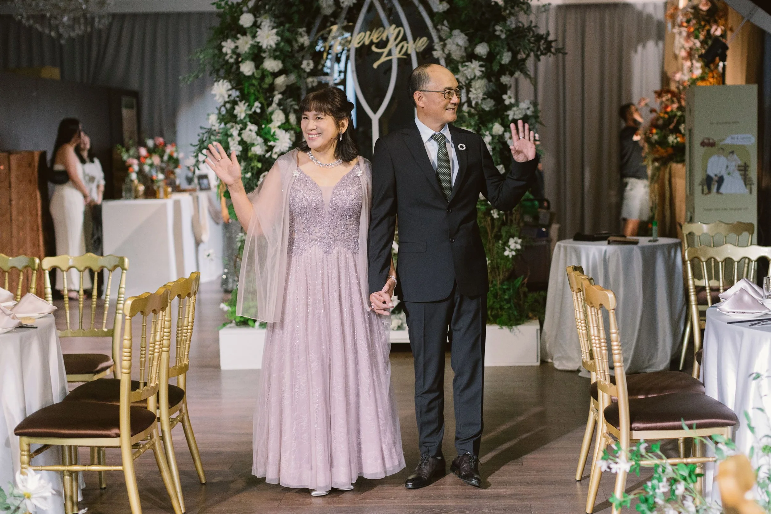 A woman in a lavender dress and a man in a black suit are smiling and waving as they walk through a decorated reception area with chairs, tables, and floral arrangements.