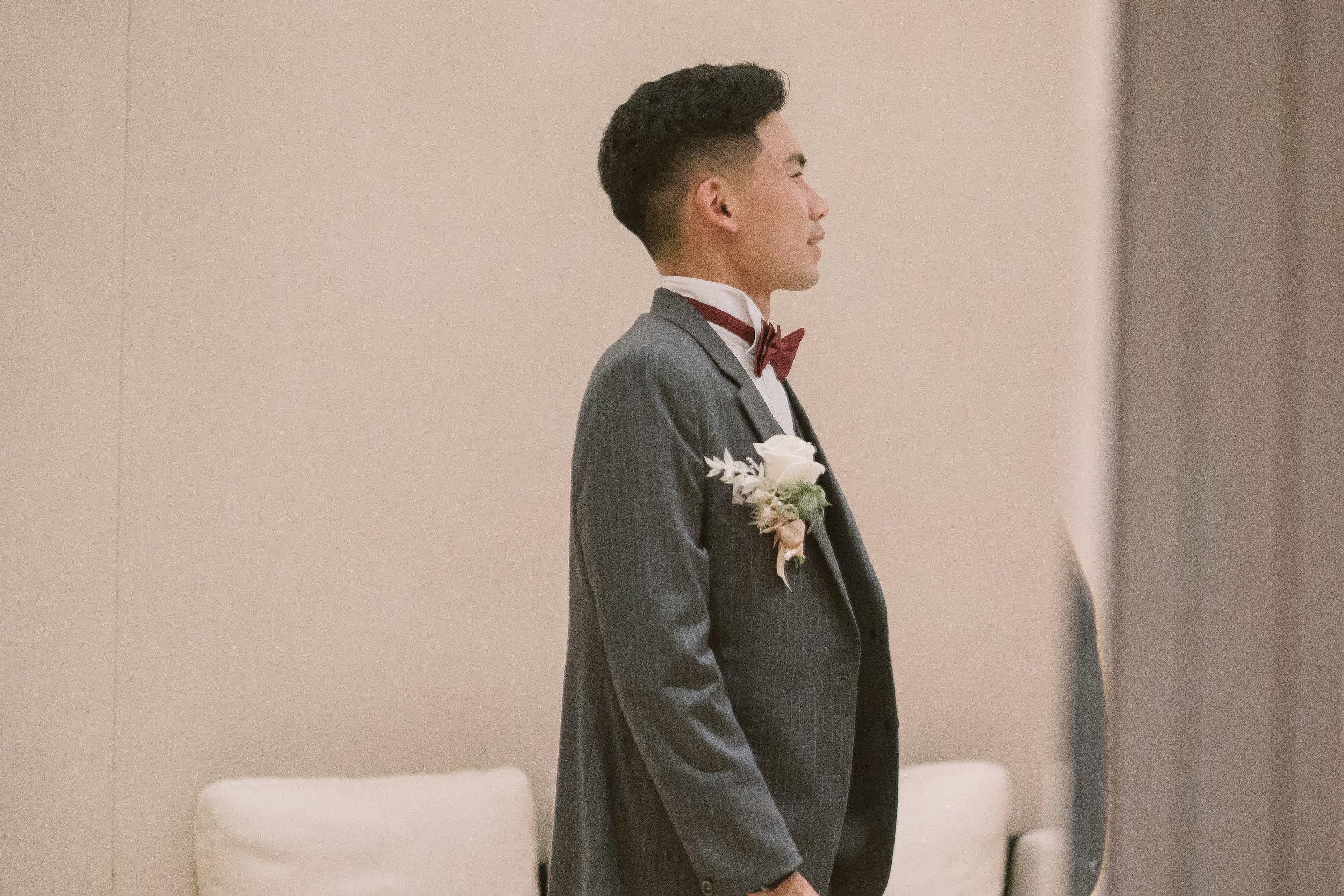 Side profile of a young man in a gray tuxedo with a burgundy bow tie and a white boutonniere, standing indoors.