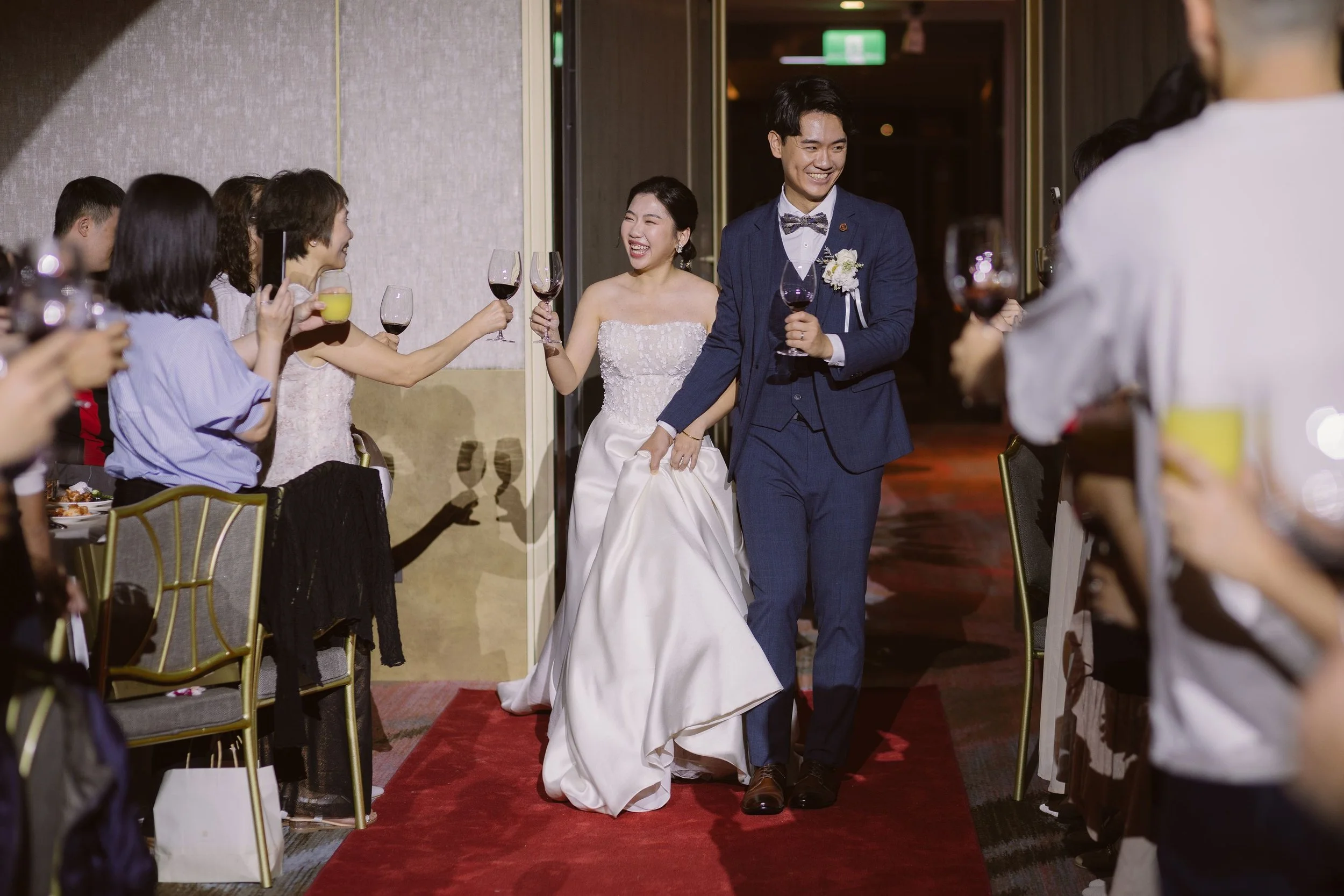 A newlywed couple walks arm-in-arm down a red carpet through a reception area, smiling and holding glasses of wine, surrounded by seated guests raising their drinks in celebration.