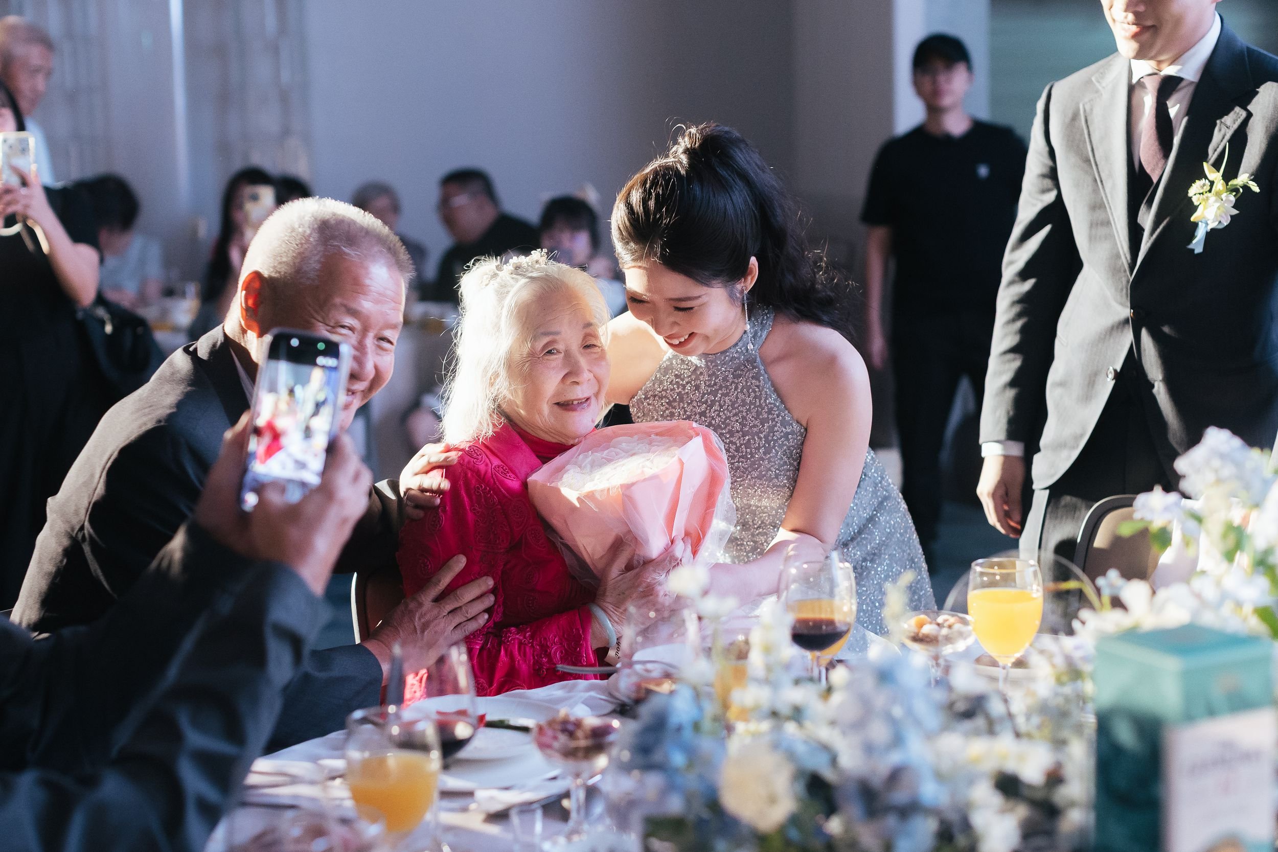 A gathering of people at a celebration, with a young woman hugging an elderly woman and people taking photos in a decorated indoor setting.