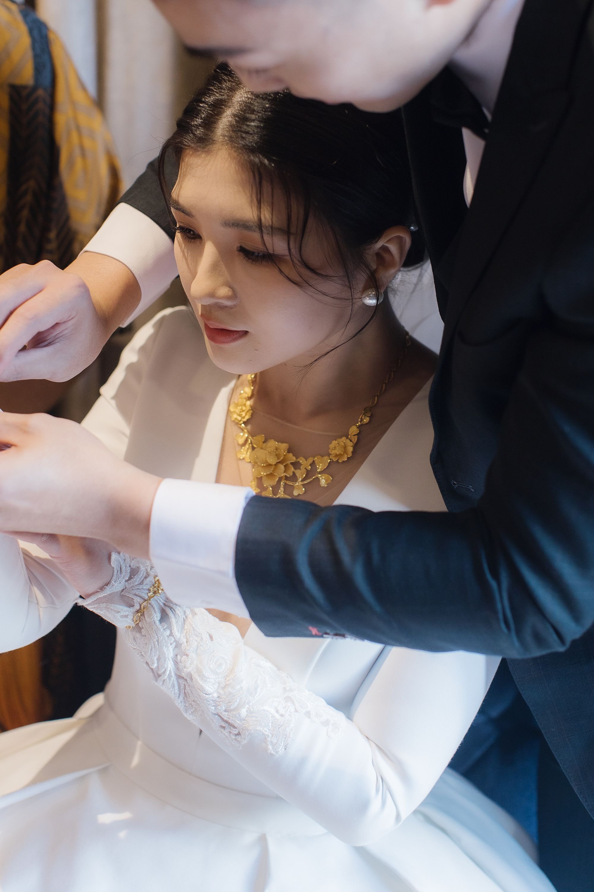 A woman in a wedding dress with lace details on the sleeves, wearing a gold floral necklace and pearl earrings, leaning forward while being assisted by a man in a black suit and white shirt.