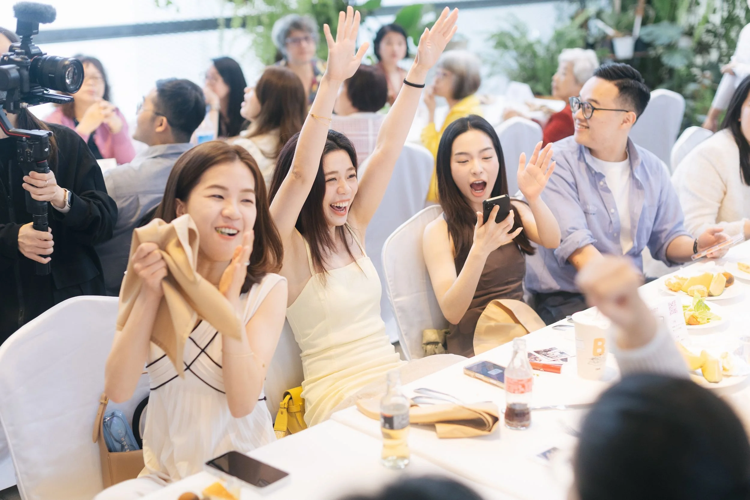 A group of young women and a young man are sitting at a table during a social event, smiling and raising their hands. There is a camera person to the left recording the event, and the table has plates of food, drinks, and smartphones.