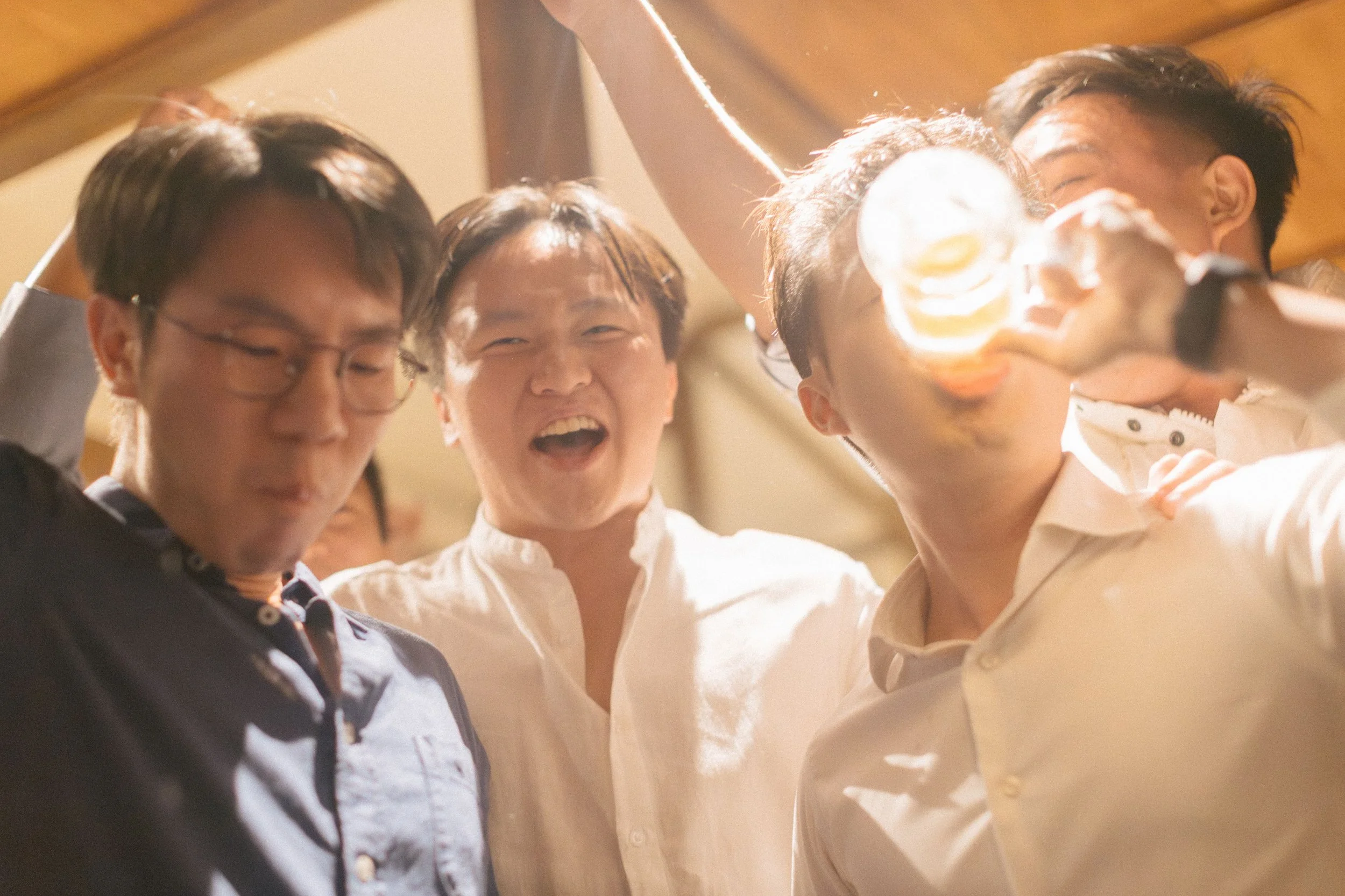 A group of four friends gathered closely, smiling, with two of them making a toast with glasses of beer.