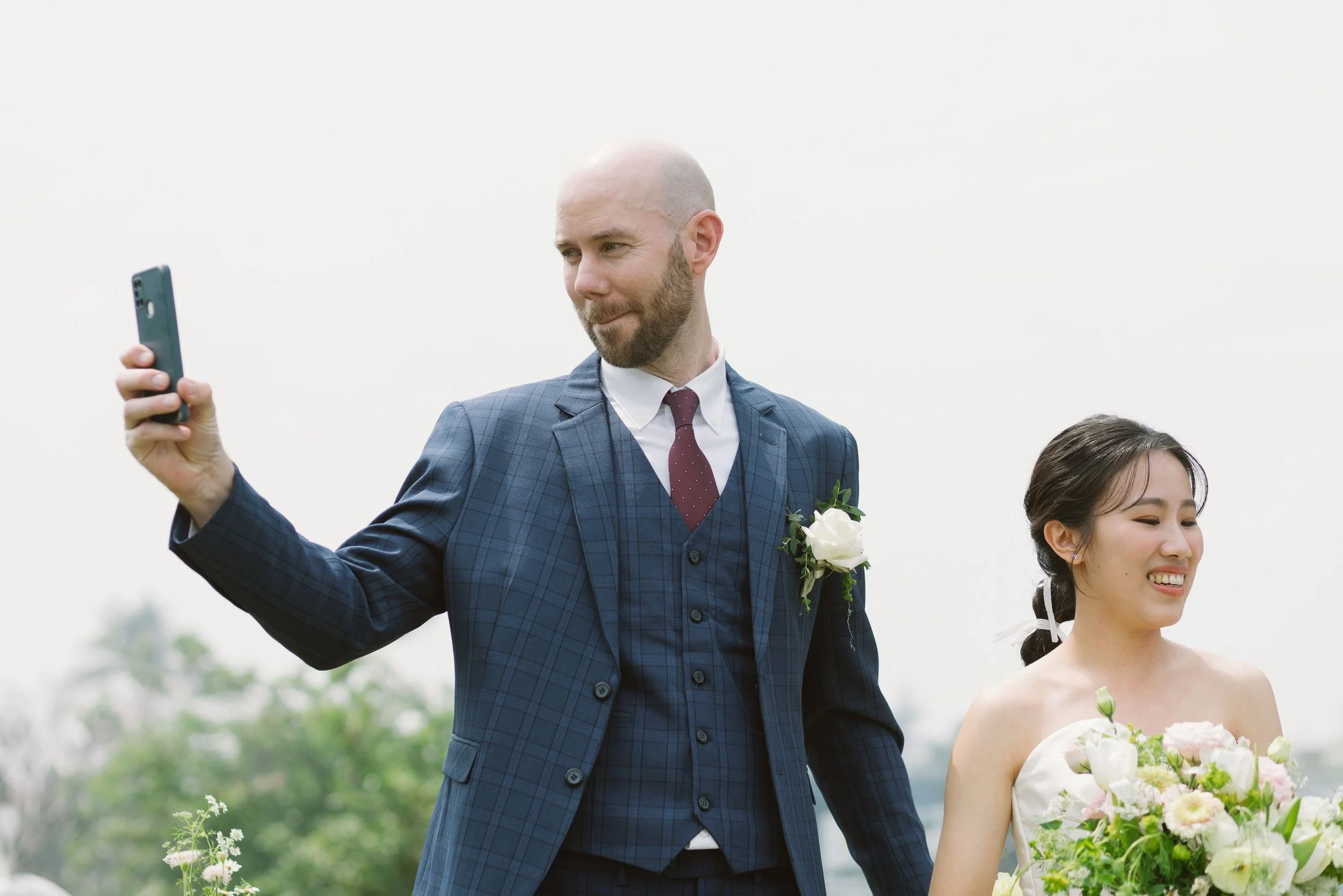 A man in a blue checkered suit taking a selfie at a wedding, with a woman holding a bouquet of flowers beside him.