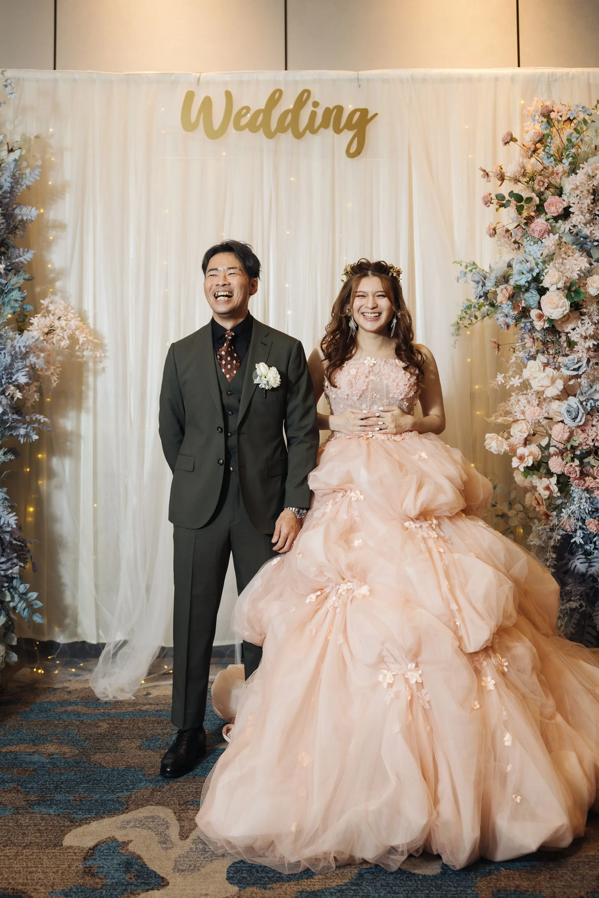A smiling bride and groom standing together at a wedding reception. The bride is wearing a pink gown with floral details, and the groom is dressed in a gray suit with a boutonniere. They are in front of a cream-colored backdrop with the word 'Wedding