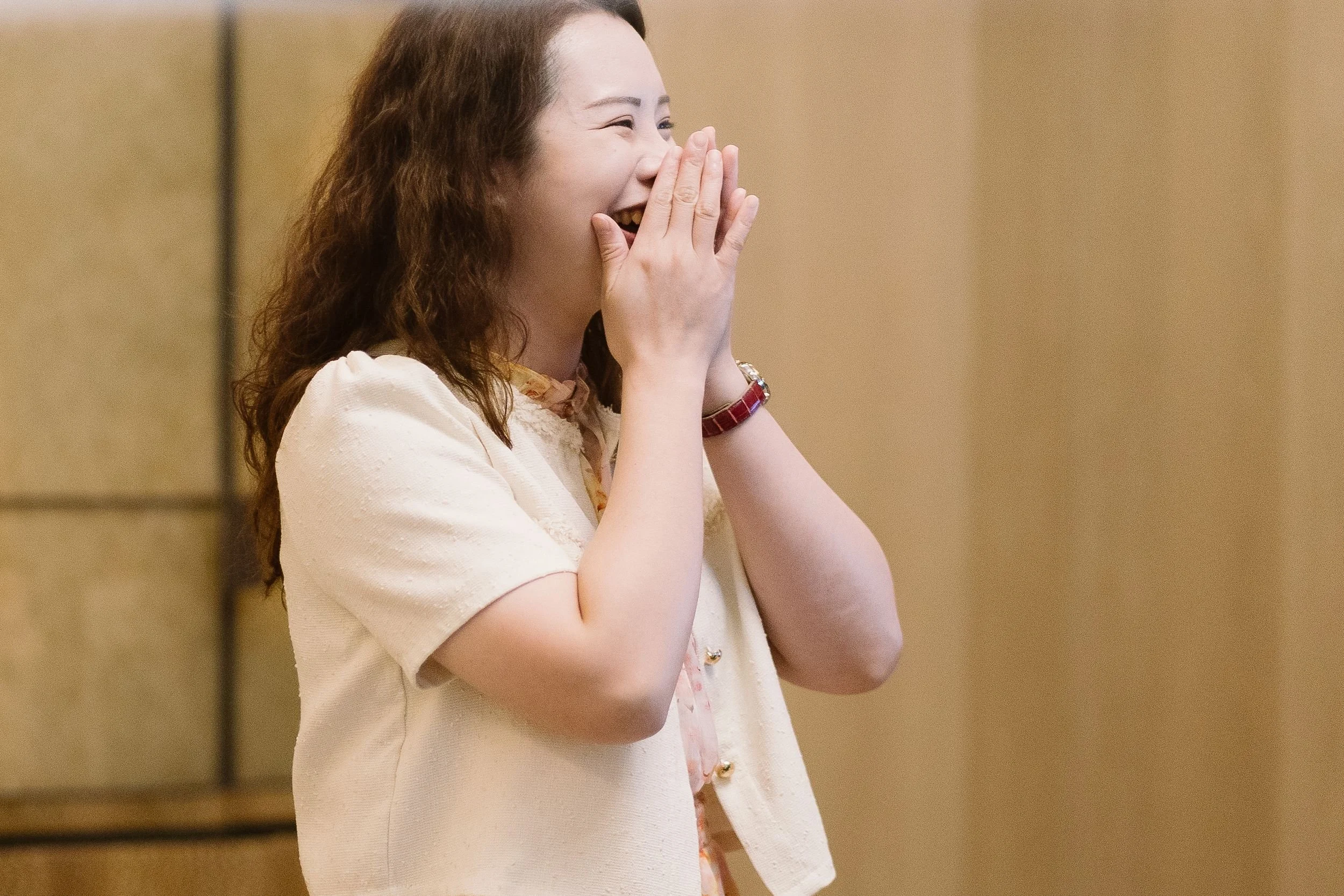 A woman with long curly hair smiling and covering her mouth with her hands, wearing a light-colored short-sleeved top and a red wristwatch, standing against a plain background.