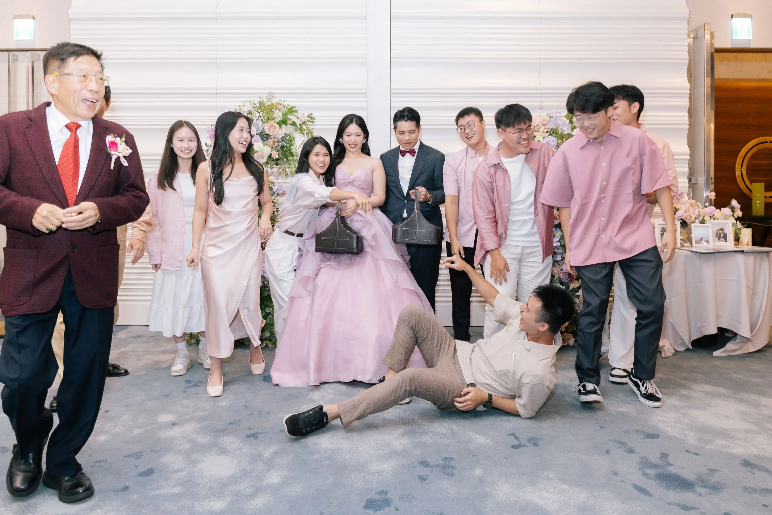 Group of people at a celebration, with a woman in a pink dress as the center, surrounded by others smiling and interacting, with floral decorations in the background.
