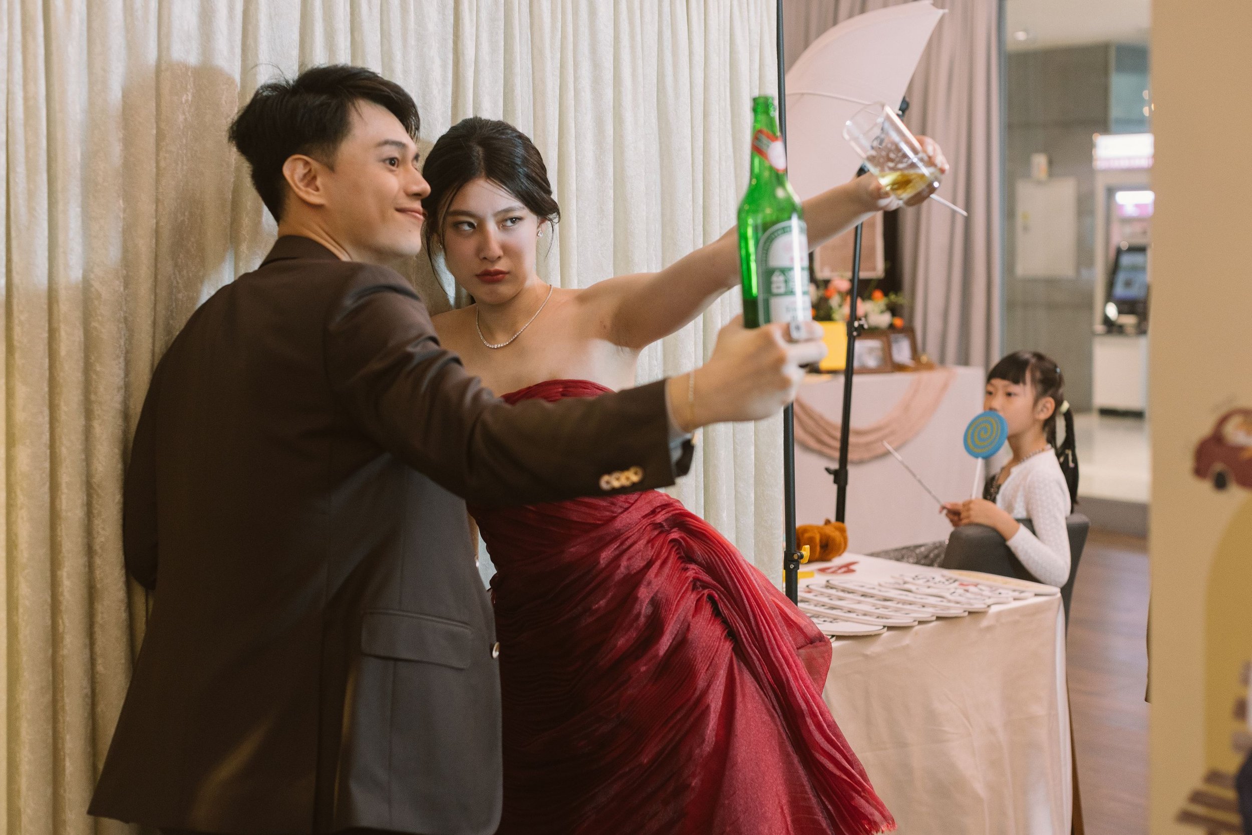 A woman in a red dress taking a selfie with a man in a dark suit at a party or event, with children and decorations in the background.